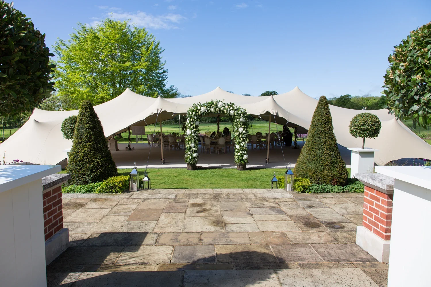 Wedding tent set up outdoors with white tent, decorated with a floral arch, surrounded by neatly trimmed bushes and trees, on a sunny day with blue sky.