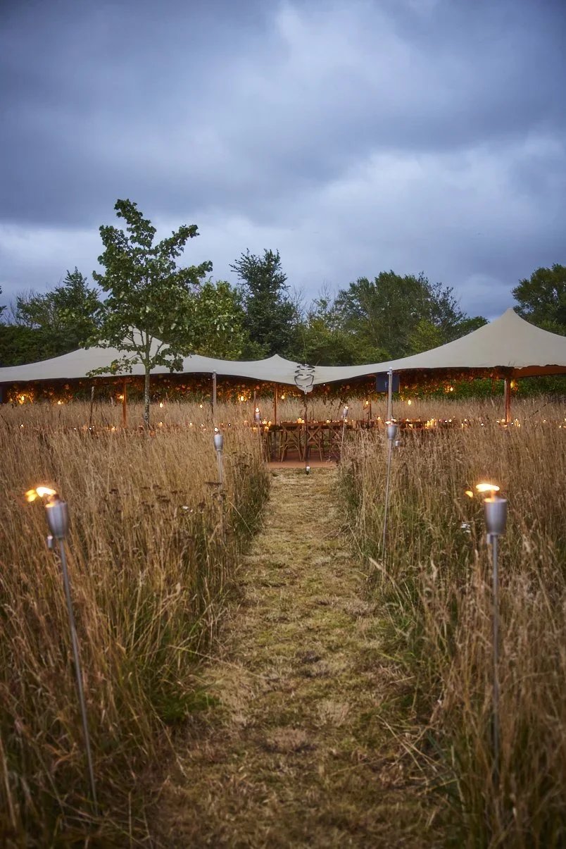 A pathway through a grassy field leading to a decorated event tent under a cloudy sky at dusk or evening.