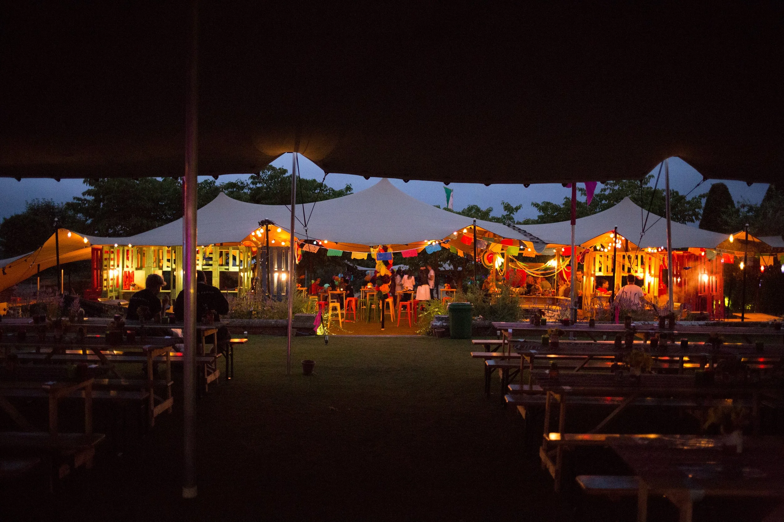 A large butterfly-shaped tent set up on a grassy field during the day with a partly cloudy sky, trees in the background, and a fence nearby. Several people are around, one holding a phone and two others wearing vests.