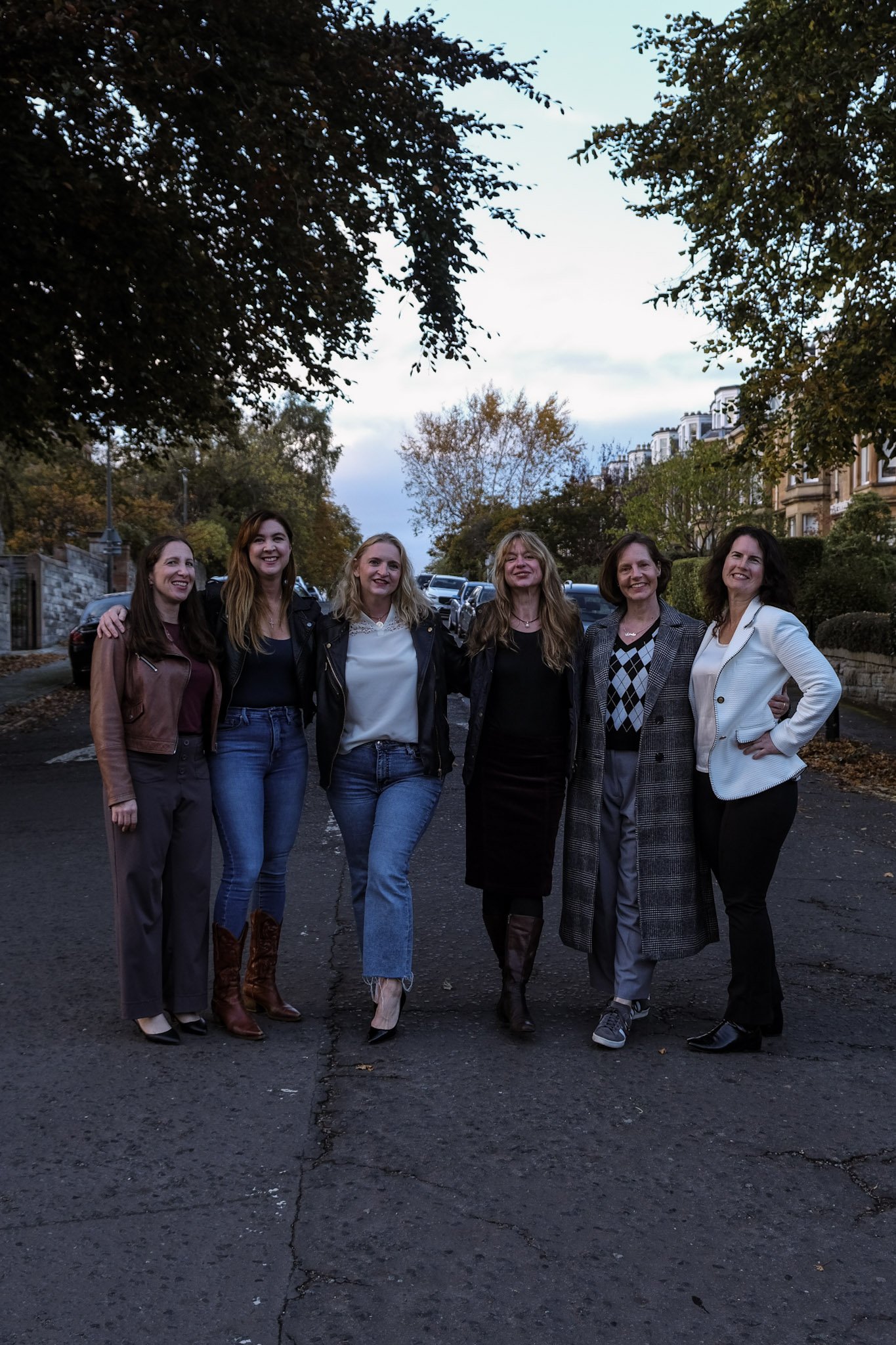 Group of six women standing arm in arm on a street at dusk, smiling and posing for the photo with trees and houses in the background.