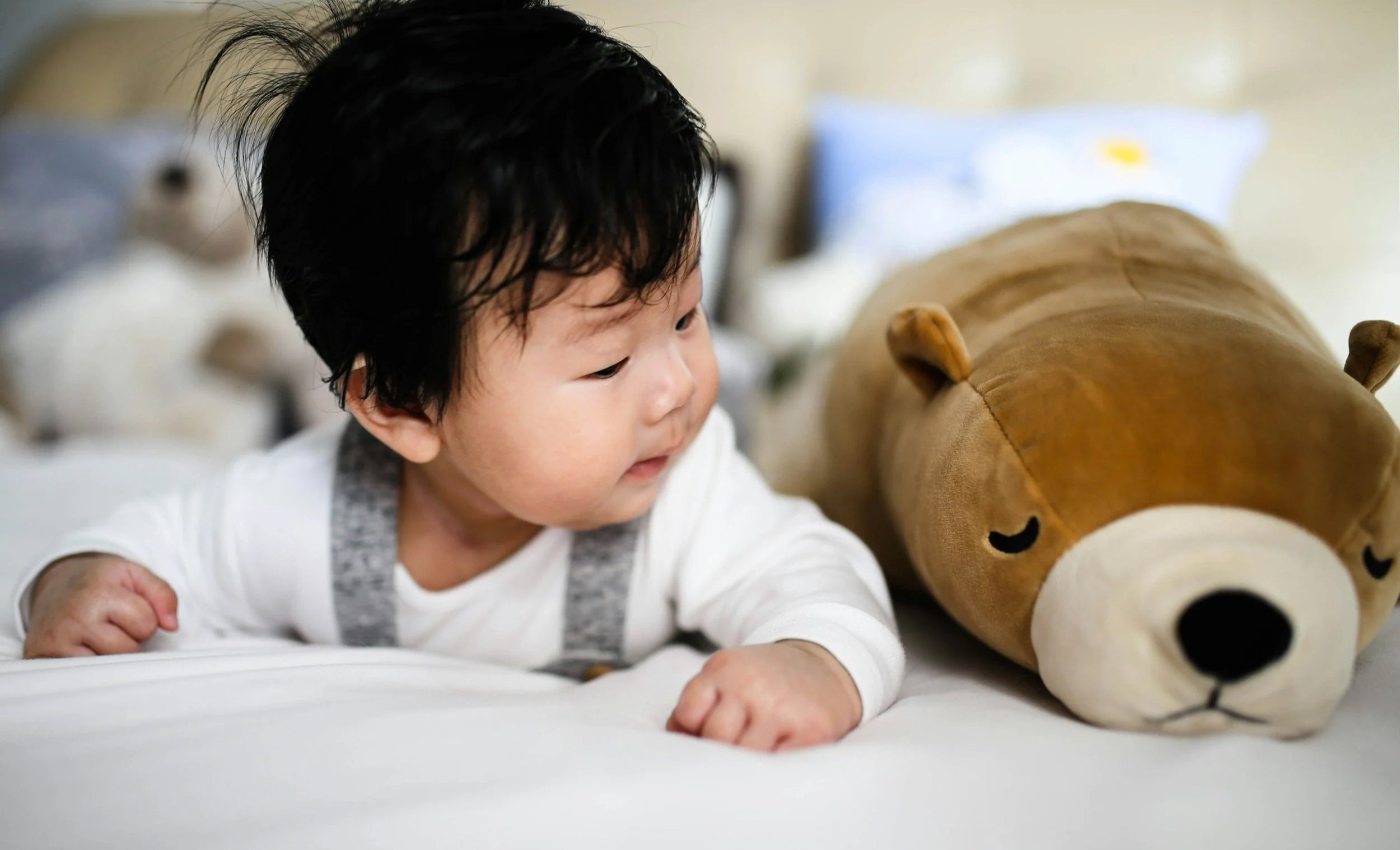 Infant with black hair lies next to big brown bear toy on a play mat