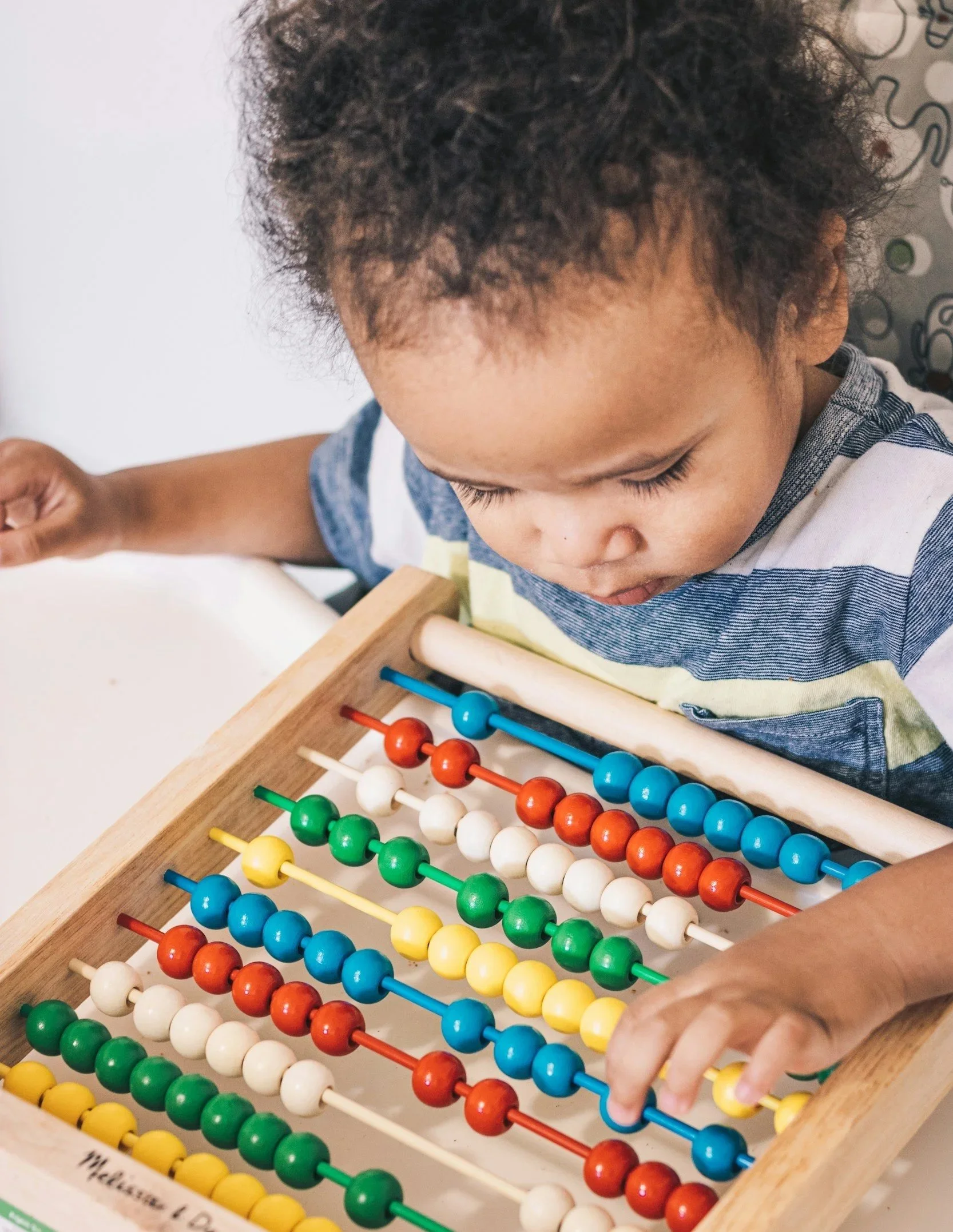 A young child with curly hair playing with a colorful wooden abacus in a Montessori classroom.