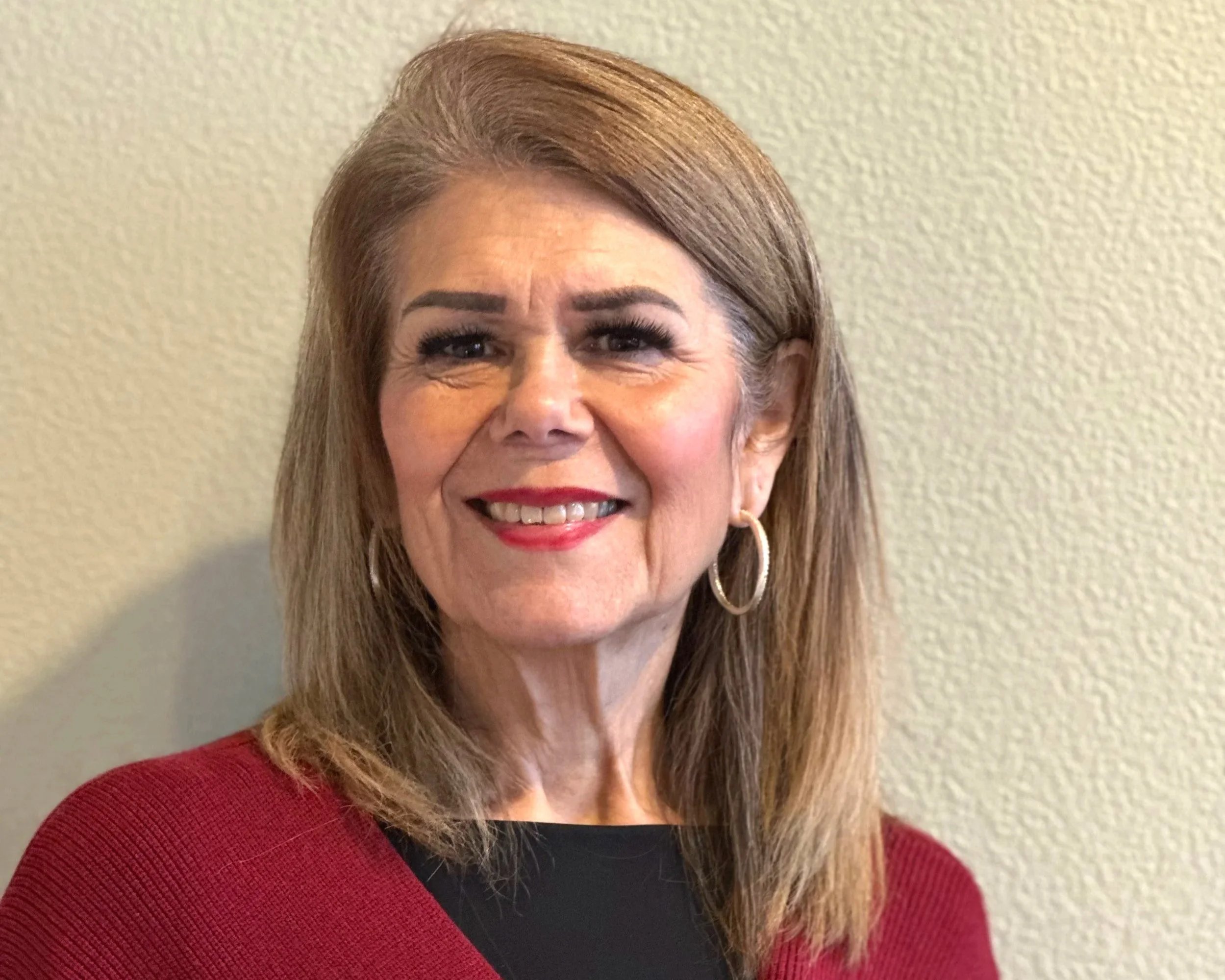 Debra Robertson, the founder and director of Montessori Davis Academy – a woman with shoulder-length, light brown hair, wearing hoop earrings, a red blazer over a black top, smiling and standing against a beige wall.