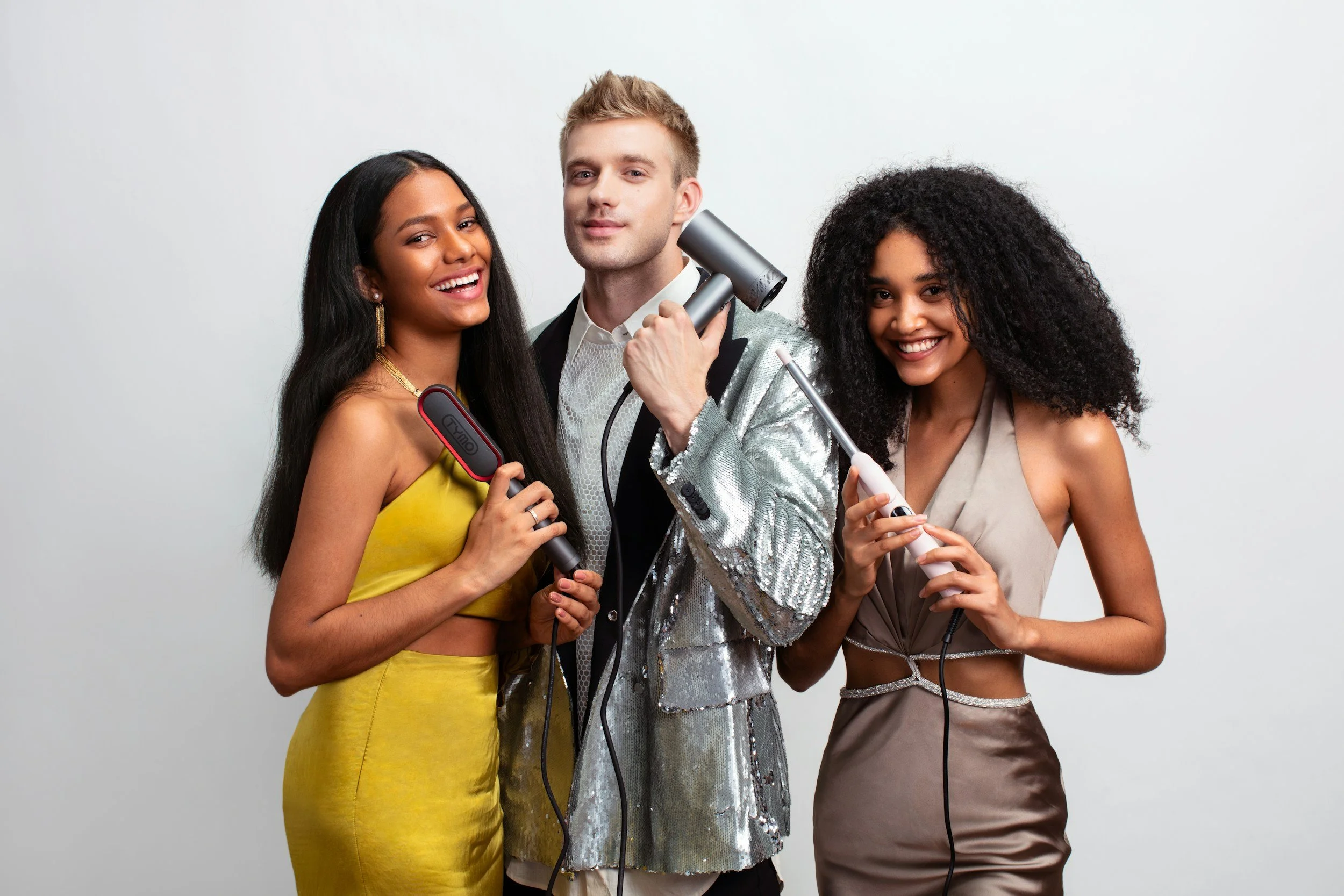 Three friends at a hair and beauty event holding hair styling tools, smiling, standing against a white background.