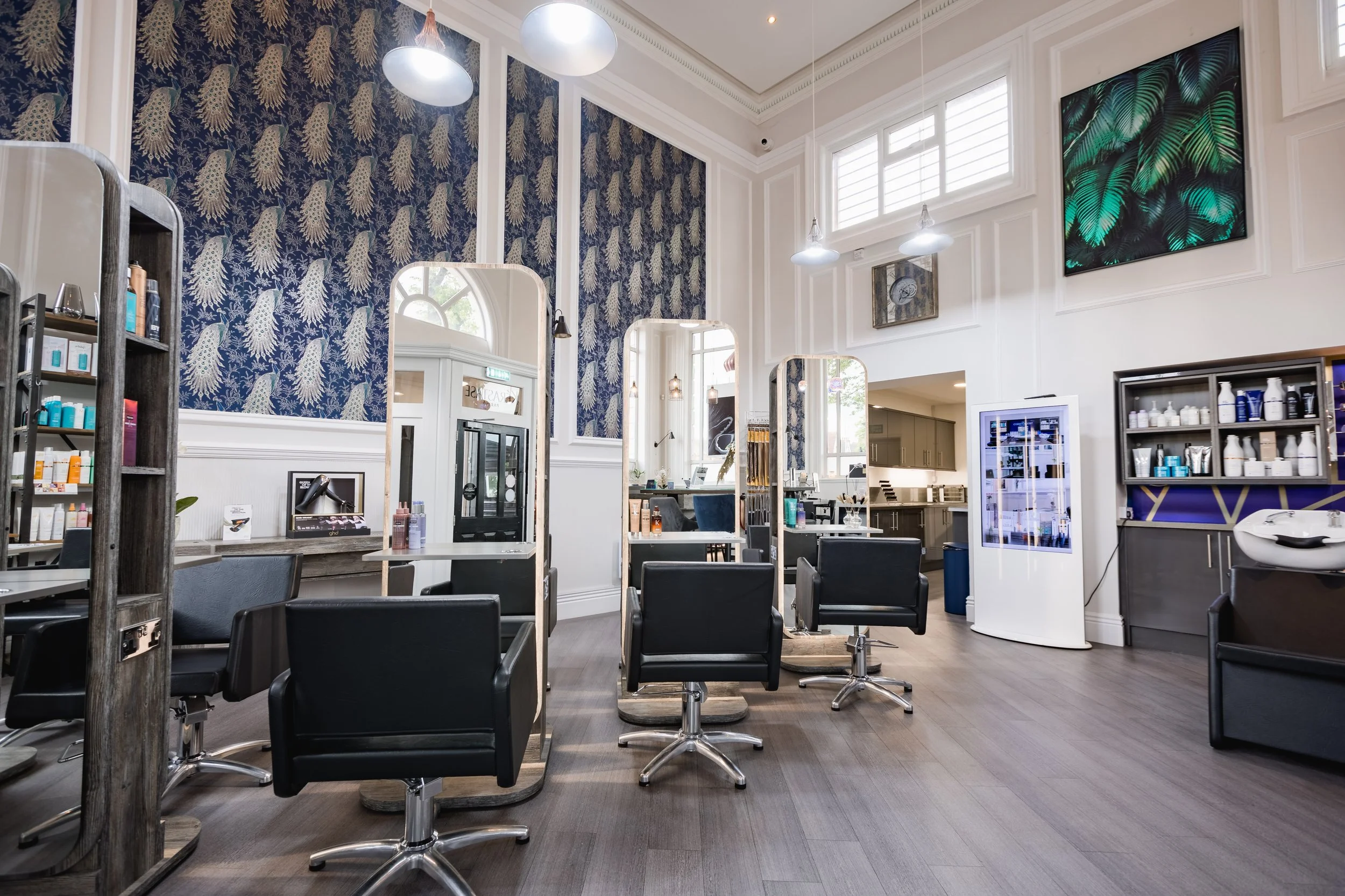 Interior of a modern hair salon with three styling stations, mirrors, salon chairs, shelves with hair products, large window, and artwork on the wall.