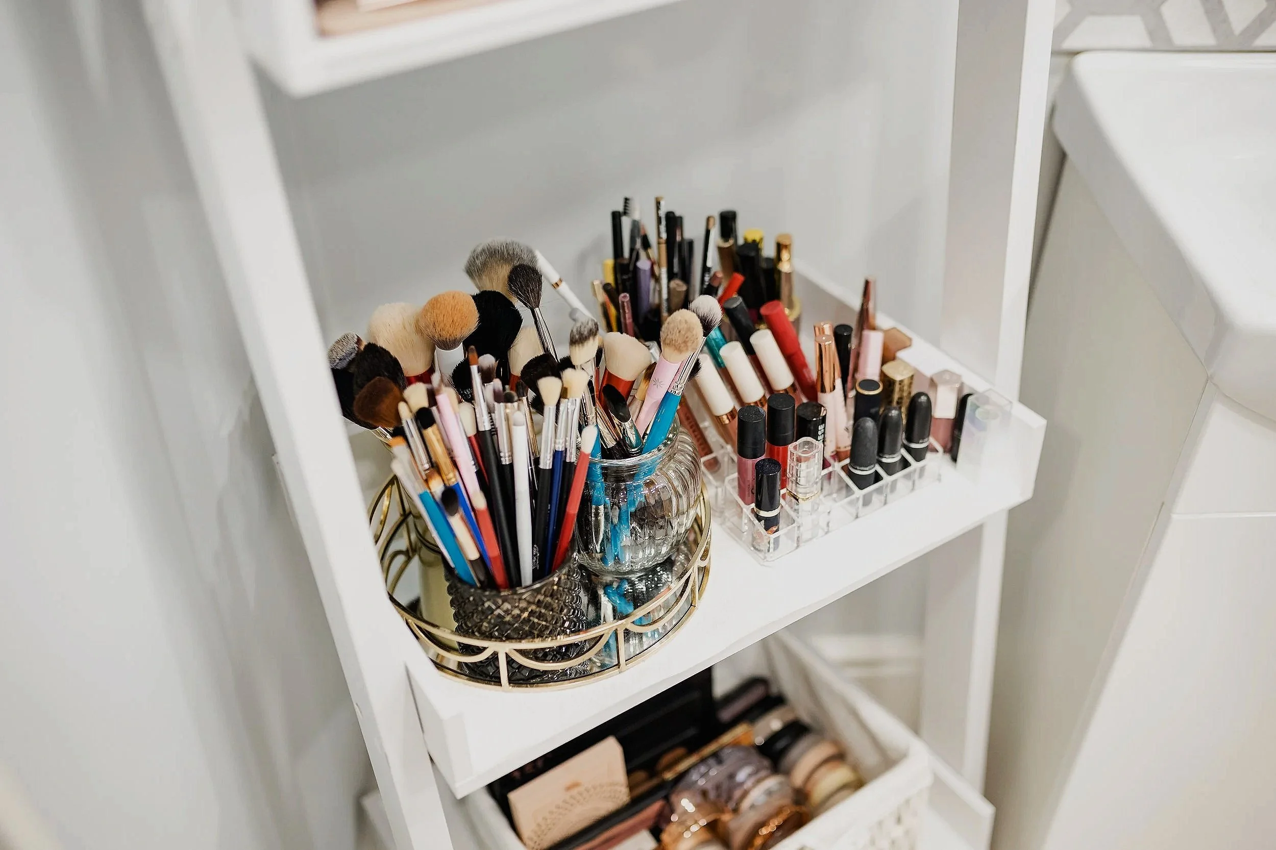 A white makeup cart with brushes, lipsticks, and makeup products organized in containers on the top shelf.