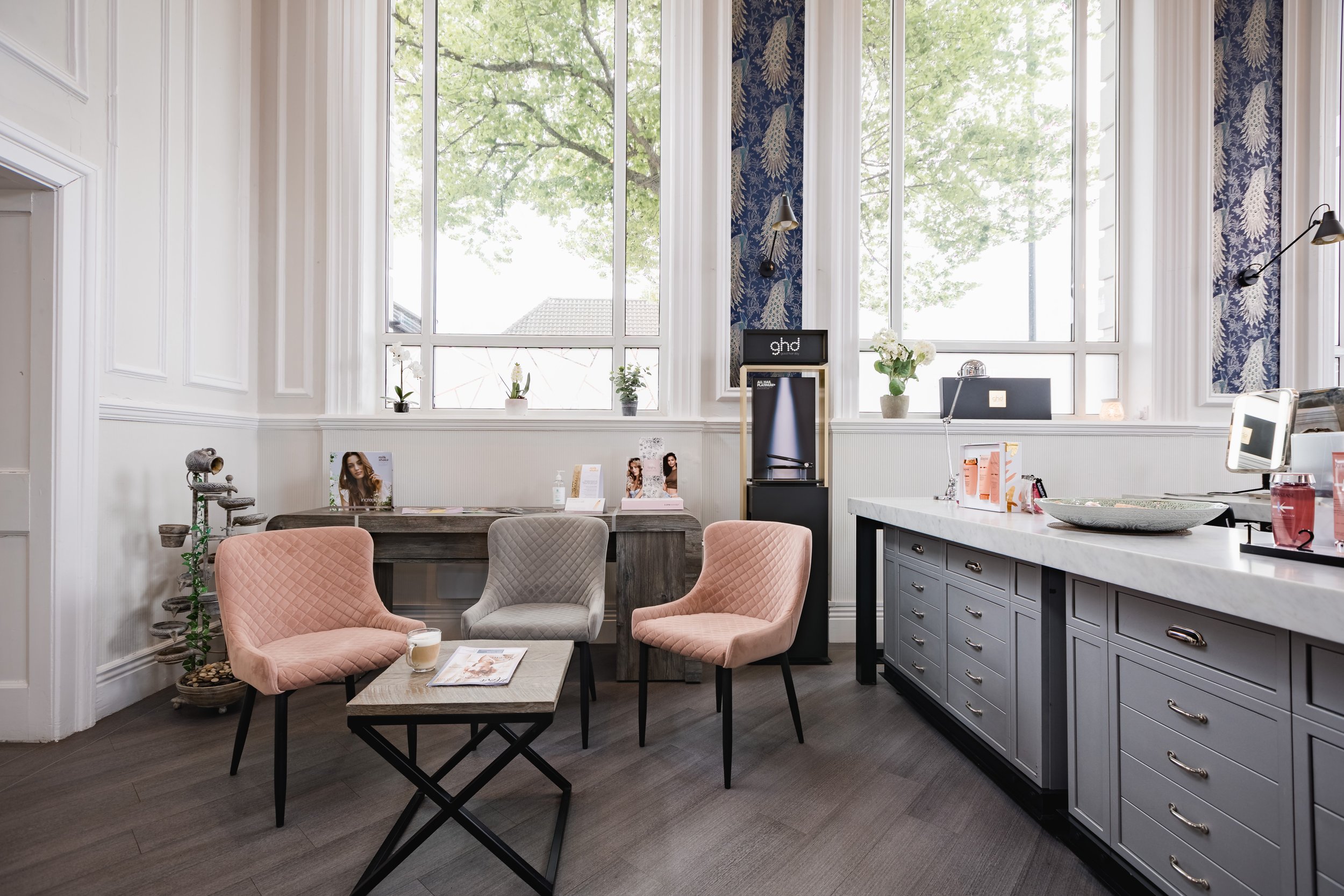 A bright interior room with tall windows, light-colored walls, and dark hardwood flooring. There are three pastel-colored chairs around a small coffee table, a wooden table with magazines and a small potted plant, and a gray cabinet with drawers and countertop. Decor includes small potted plants on the windowsill and a patterned blue curtain on one side.