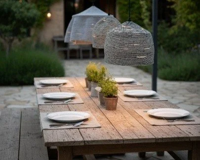 Ein Gartentisch mit sechs weißen Tellern, einer Pflanze in einem Topf, einem großen Sonnenschirm und zwei hängenden Lichter im Garten bei Sonnenuntergang.