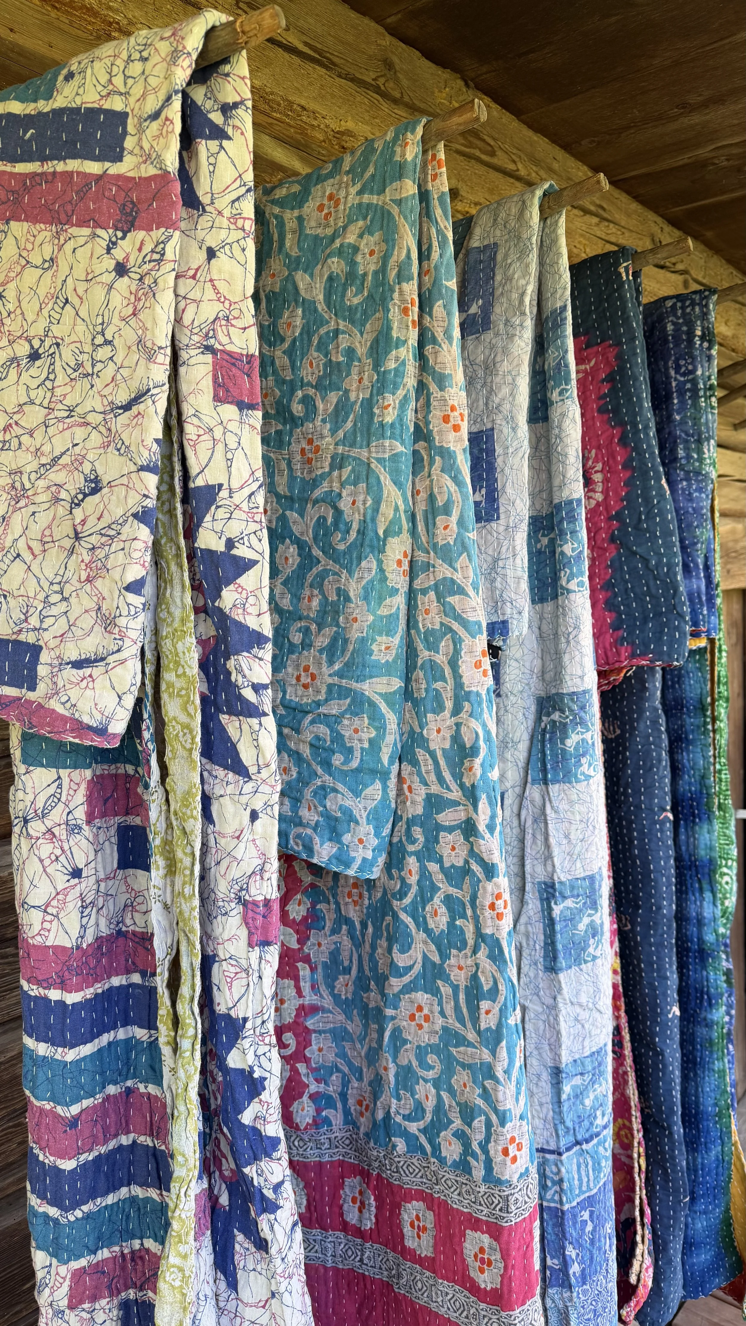 Bunte, bestickte Quilt-Decken hängen an einer Holzstange unter einer Holzdecke.