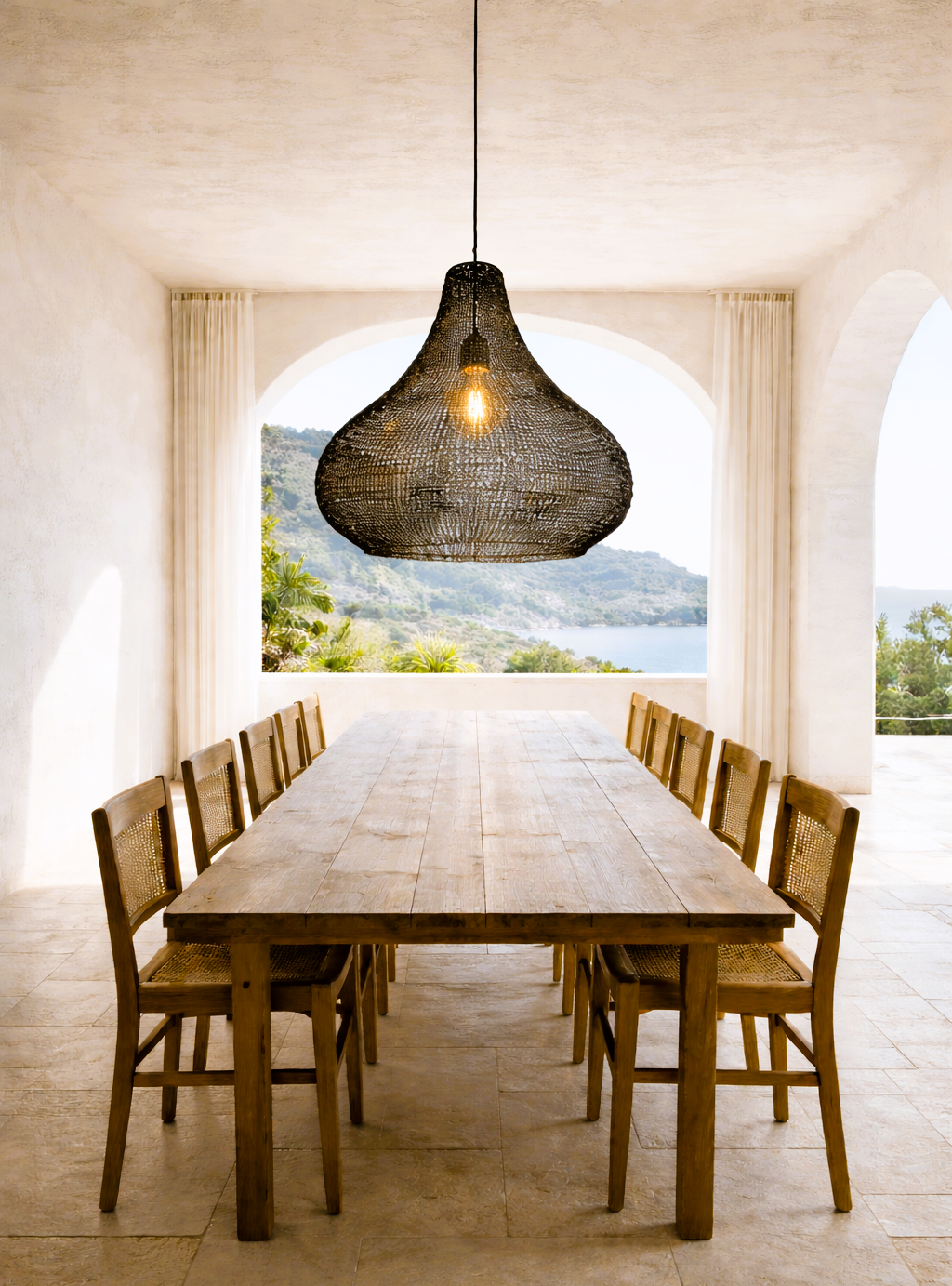 Ein heller Esszimmerraum mit einem großen Holztisch und acht Stühlen, vor einem großen Fenster mit Blick auf eine Landschaft mit Wasser und Bergen. Über dem Tisch hängt eine große, schwarze Hängeleuchte.