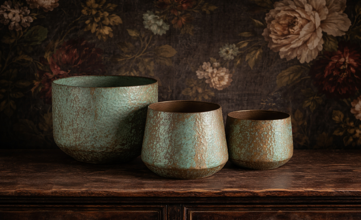 Drei handgefertigte Keramikvasen mit einer Patina in Grün, Braun und Türkis, die auf einem dunklen Holztisch vor einem floral gemusterten Hintergrund stehen.