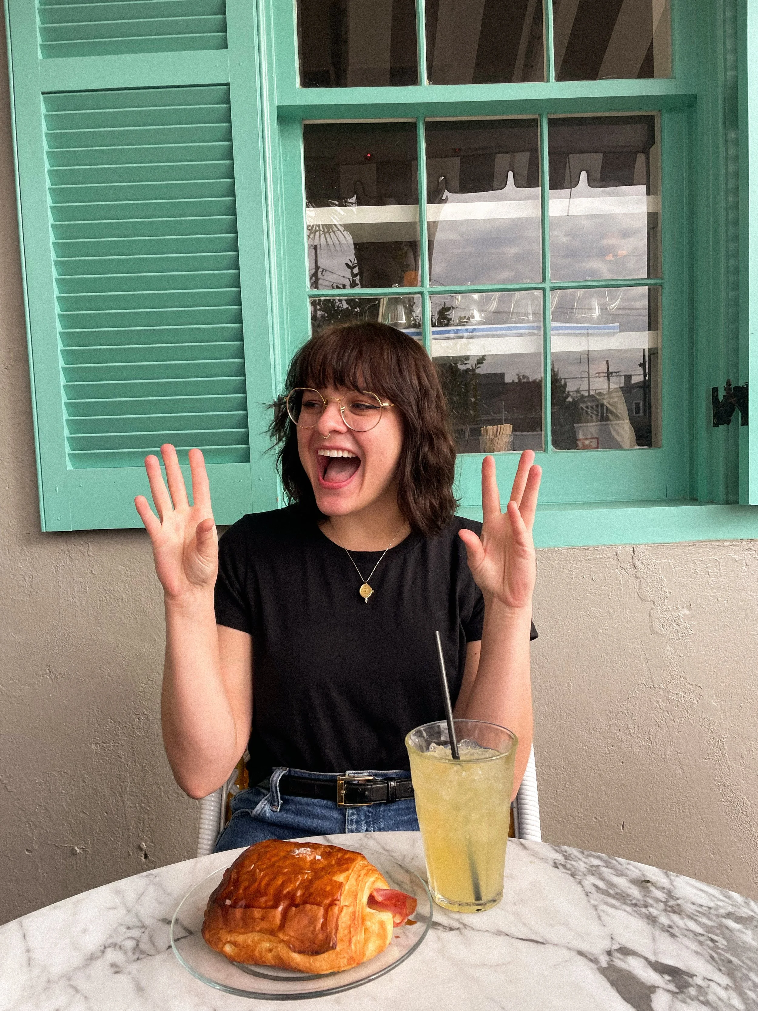 Woman with glasses smiling and raising her hands at a table with a pastry and a yellow beverage in front of a turquoise window.