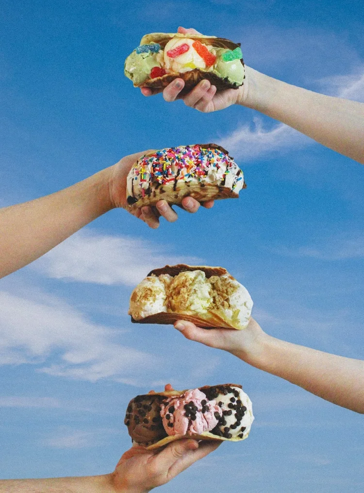 Four hands holding different ice cream sandwiches against a blue sky with clouds.