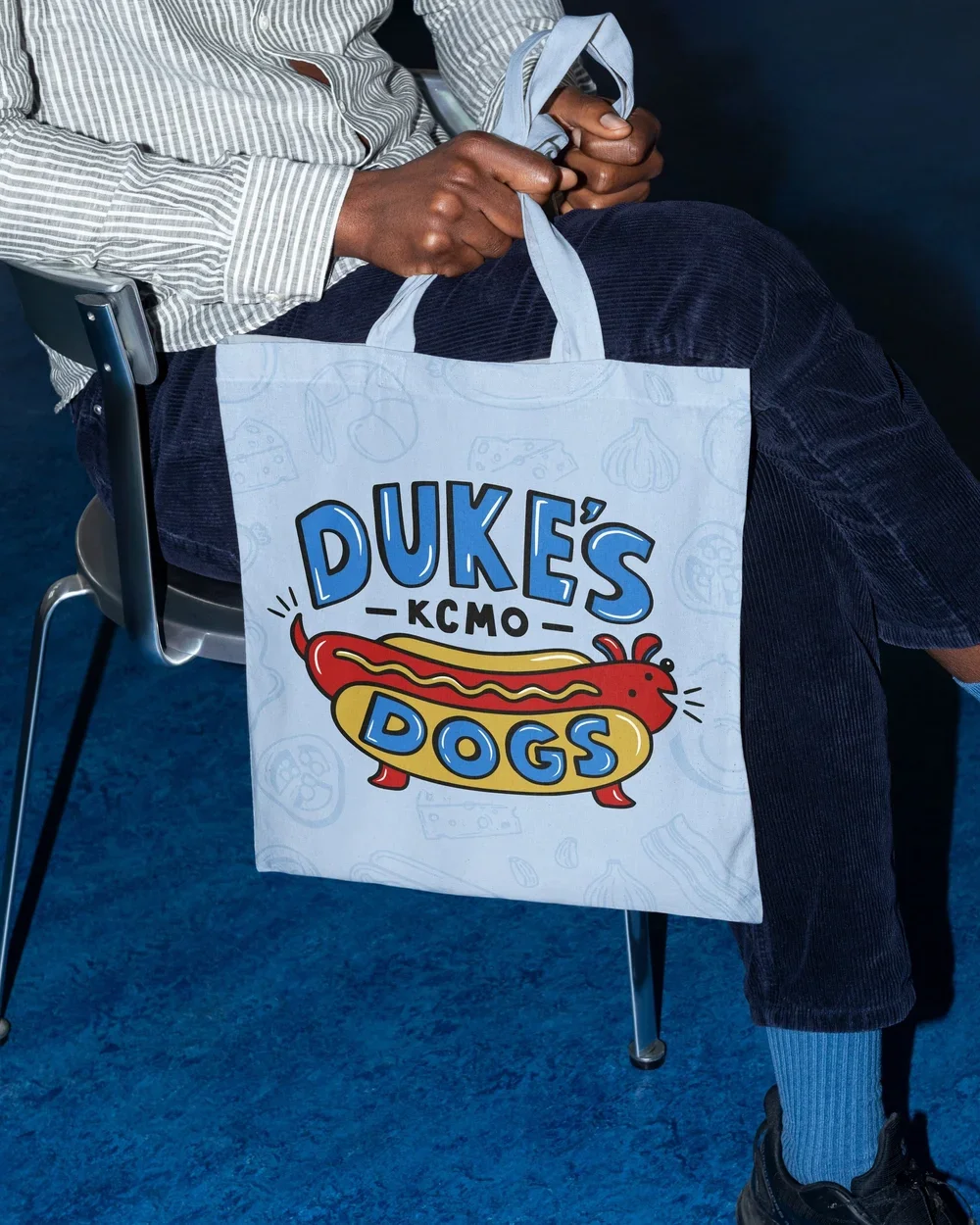 A person sitting on a chair holding a white tote bag with the logo 'Duke's Dogs' featuring a cartoon hot dog, at a KCMO (Kansas City Missouri) event or location.