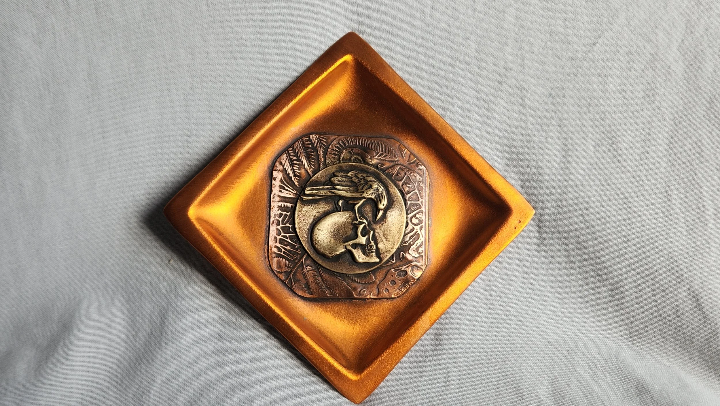 Square copper dish with a die-struck brass image of a raven atop a skull.