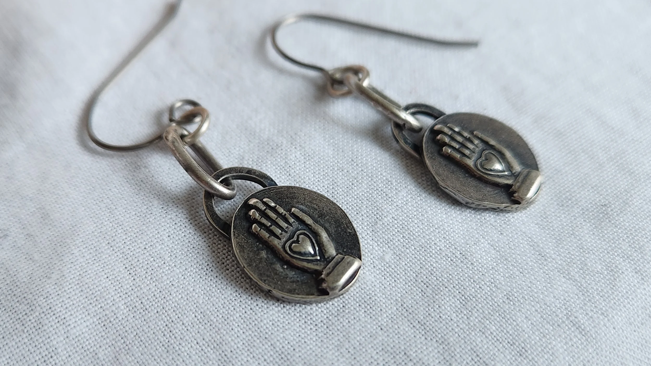 Die-struck sterling silver drop earrings with heart in hand motif and oxidized finish.