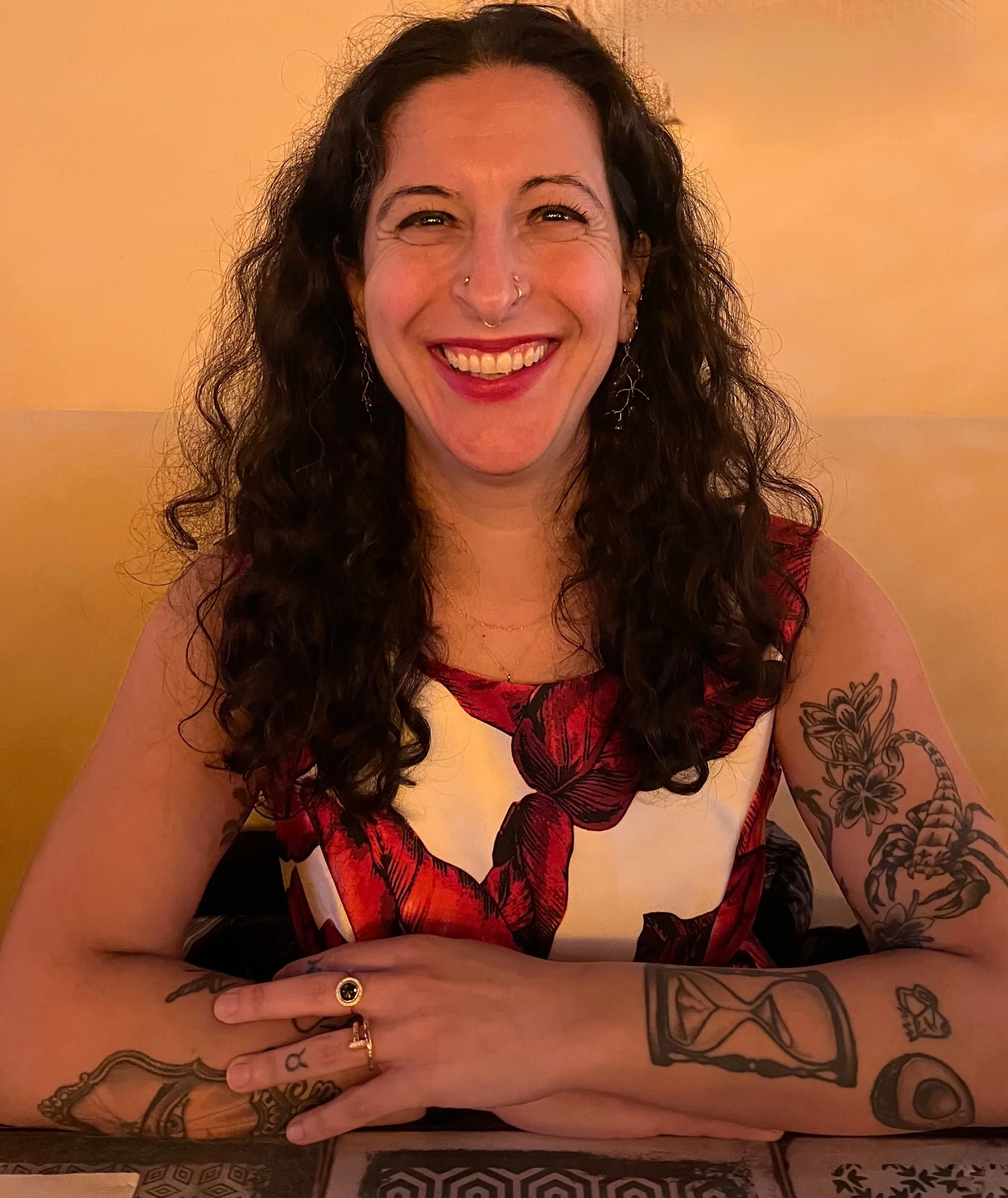 A woman with long, curly dark hair, smiling, seated at a table with tattoos on her arms, wearing a sleeveless dress with red and white floral pattern.