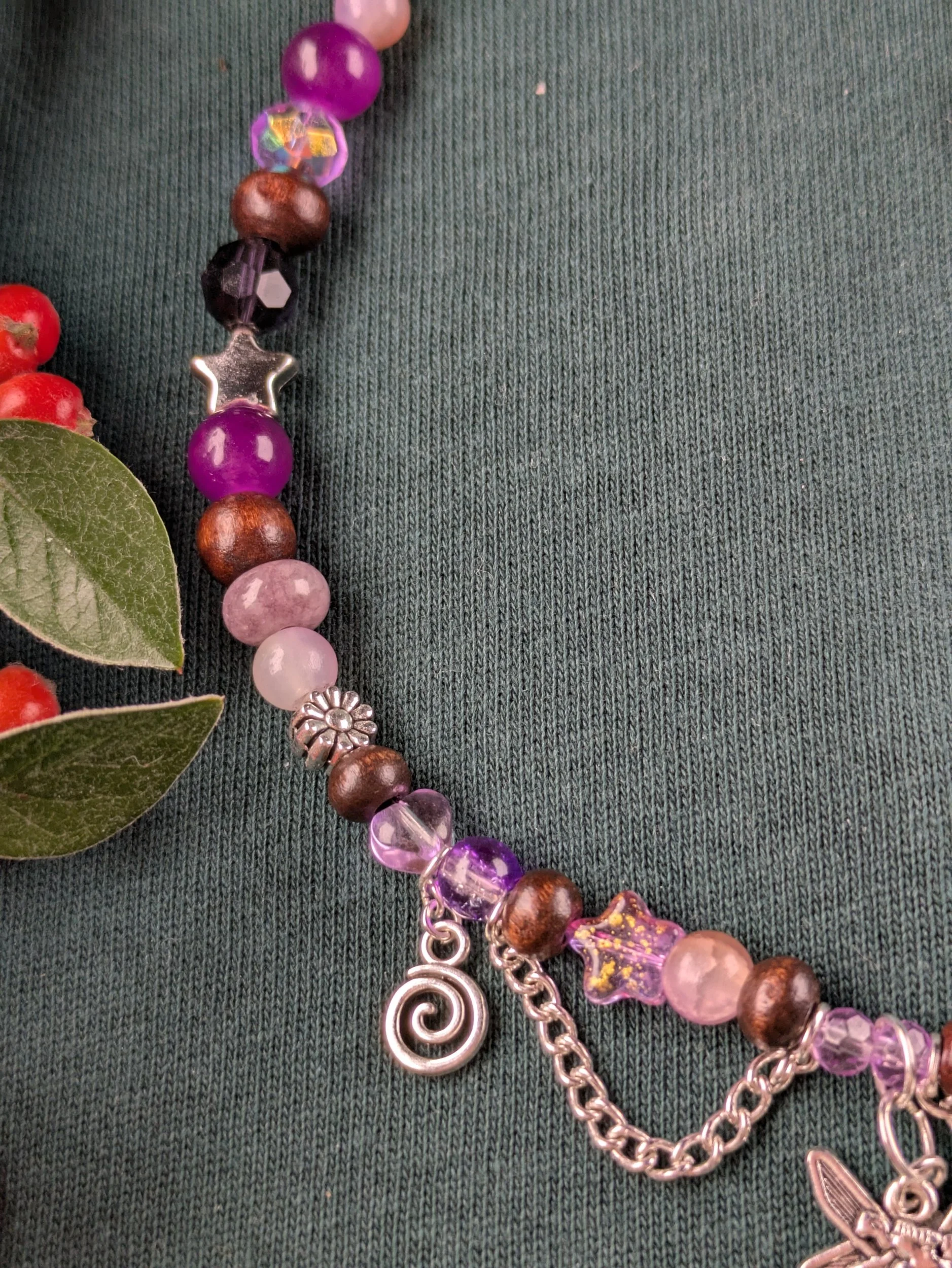 Close-up of a colorful beaded bracelet with charms on a dark green fabric background, with a sprig of red berries and green leaves on the left.