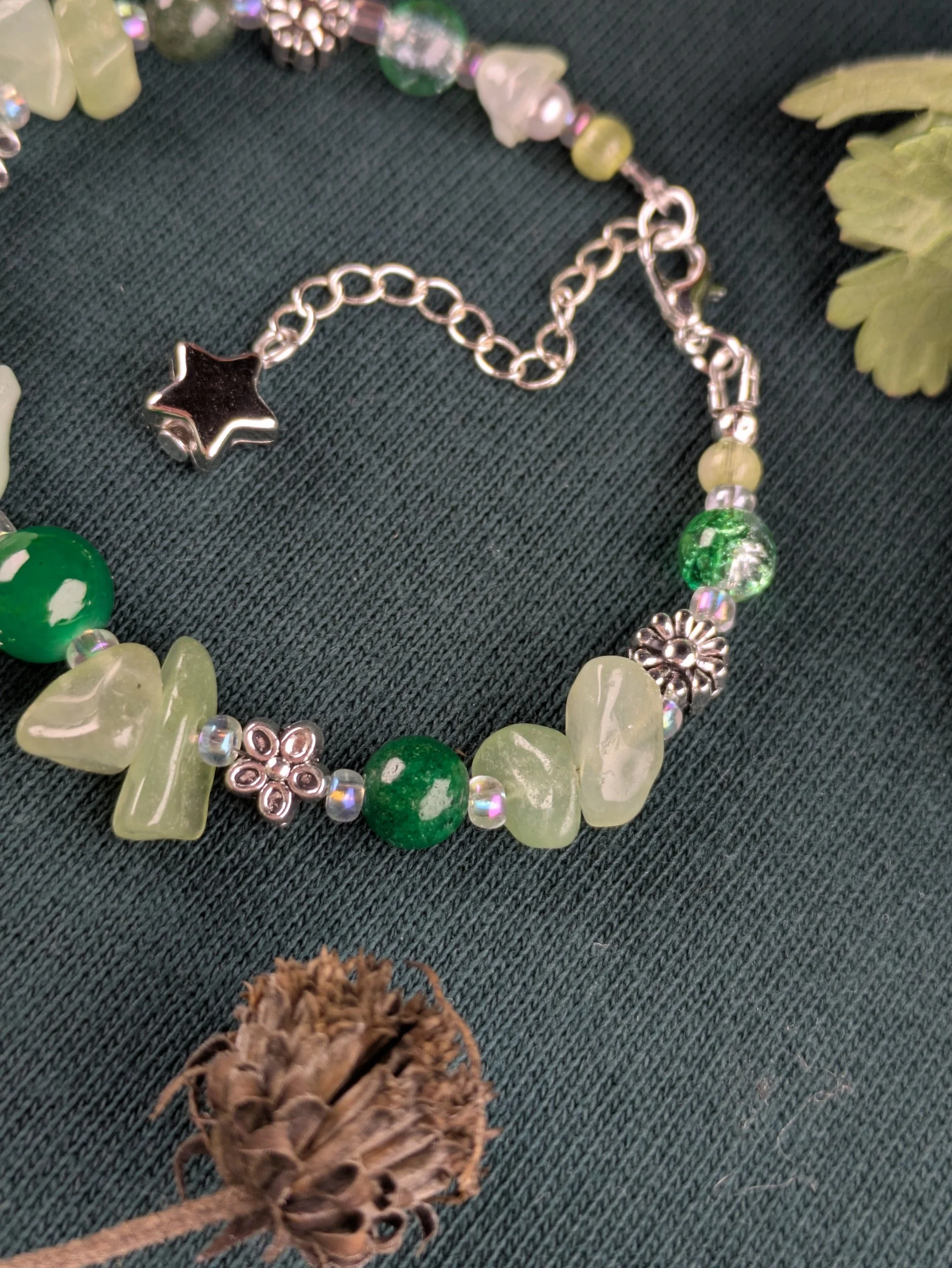 A close-up of a beaded bracelet and necklace with green, white, and silver beads, decorative metallic flower and star charms, on dark fabric. There are also some leaves and a dried seed pod nearby.