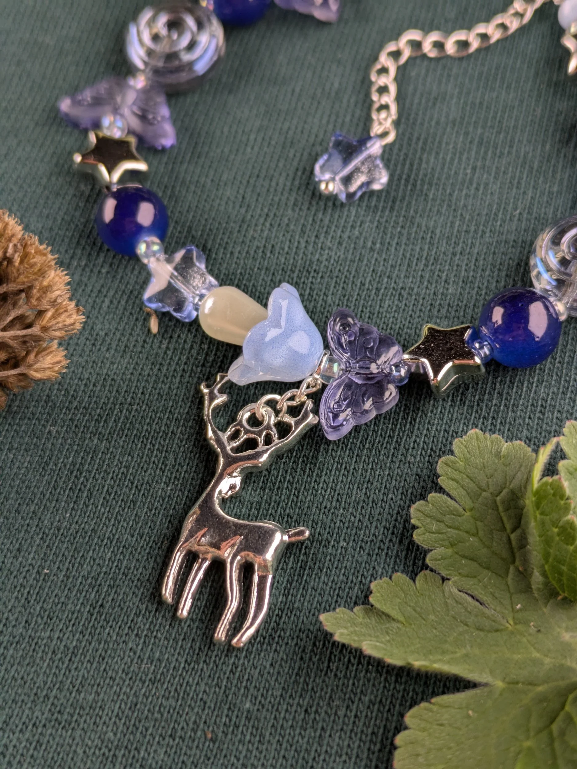 A necklace with various colored beads, including star shapes, heart shapes, and a reindeer pendant, laid on a green fabric with leaves and a pine cone nearby.