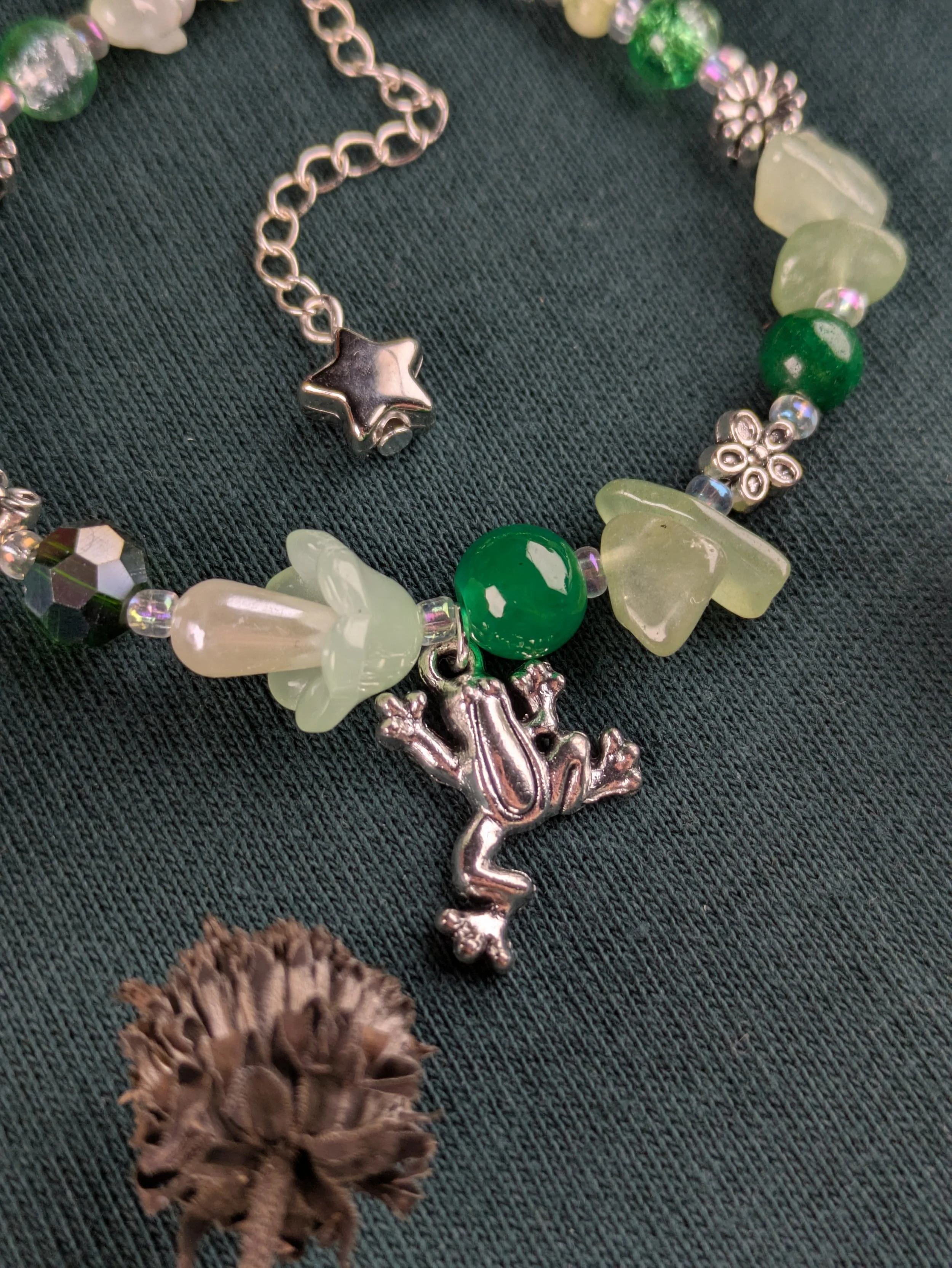 Close-up of a colorful beaded necklace with a silver frog pendant and a star-shaped clasp on a dark textured fabric background.