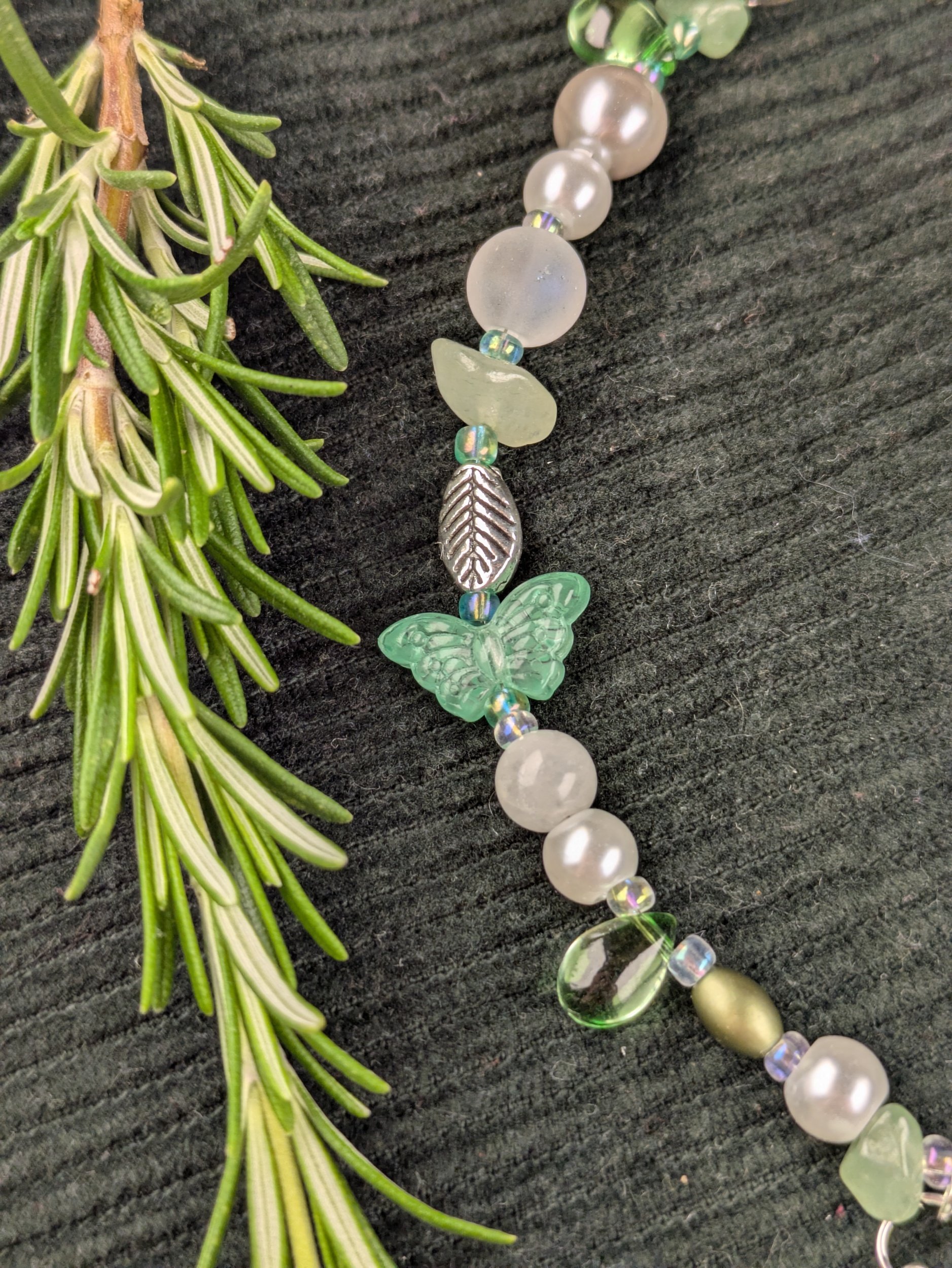 A close-up of a beaded necklace with a green butterfly, leaf-shaped, and round beads, displayed on a dark textured surface next to a sprig of green herbs.