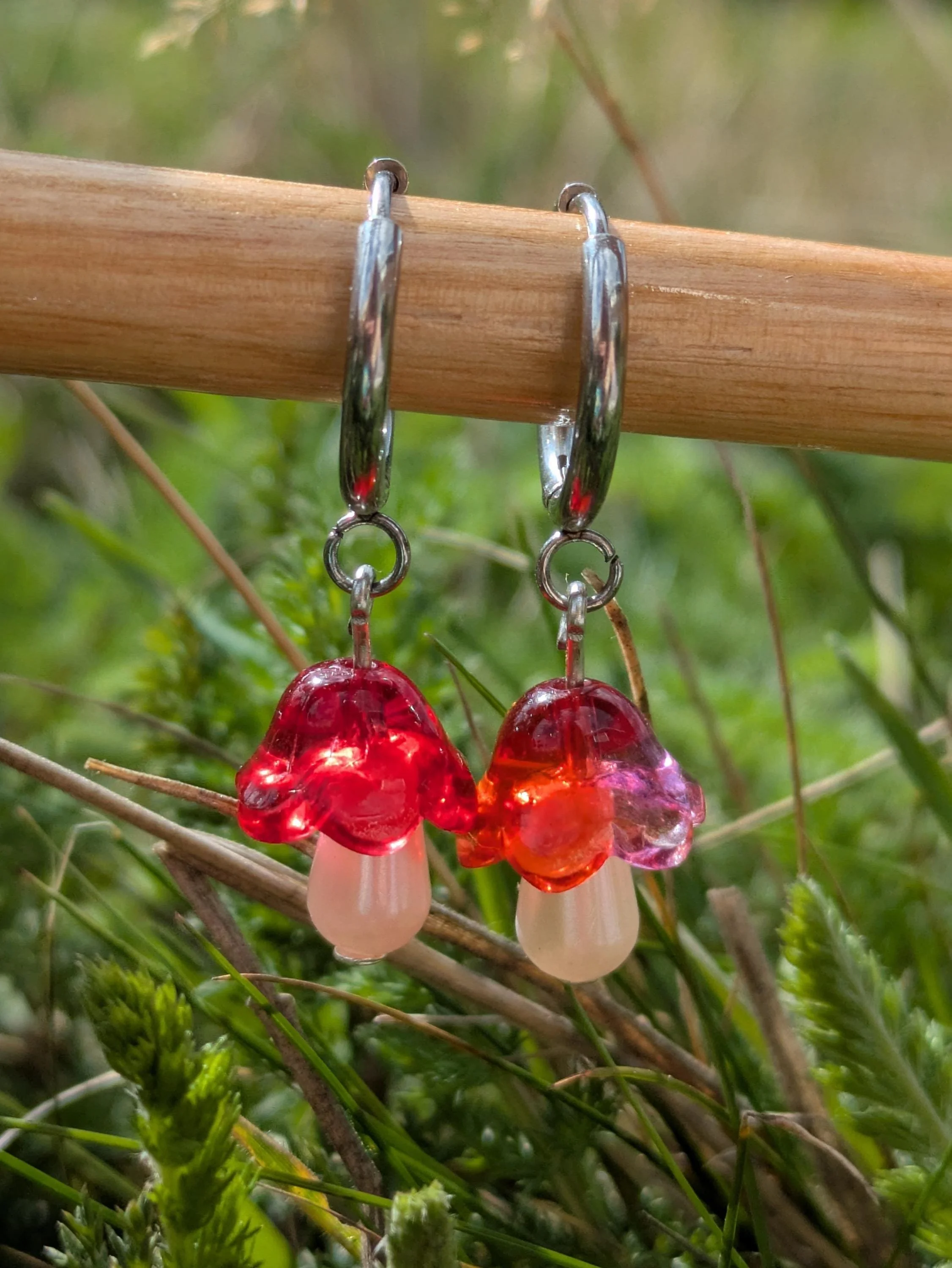 Close-up of glass flower-shaped earrings hanging from a wooden stick, with a natural grassy background.