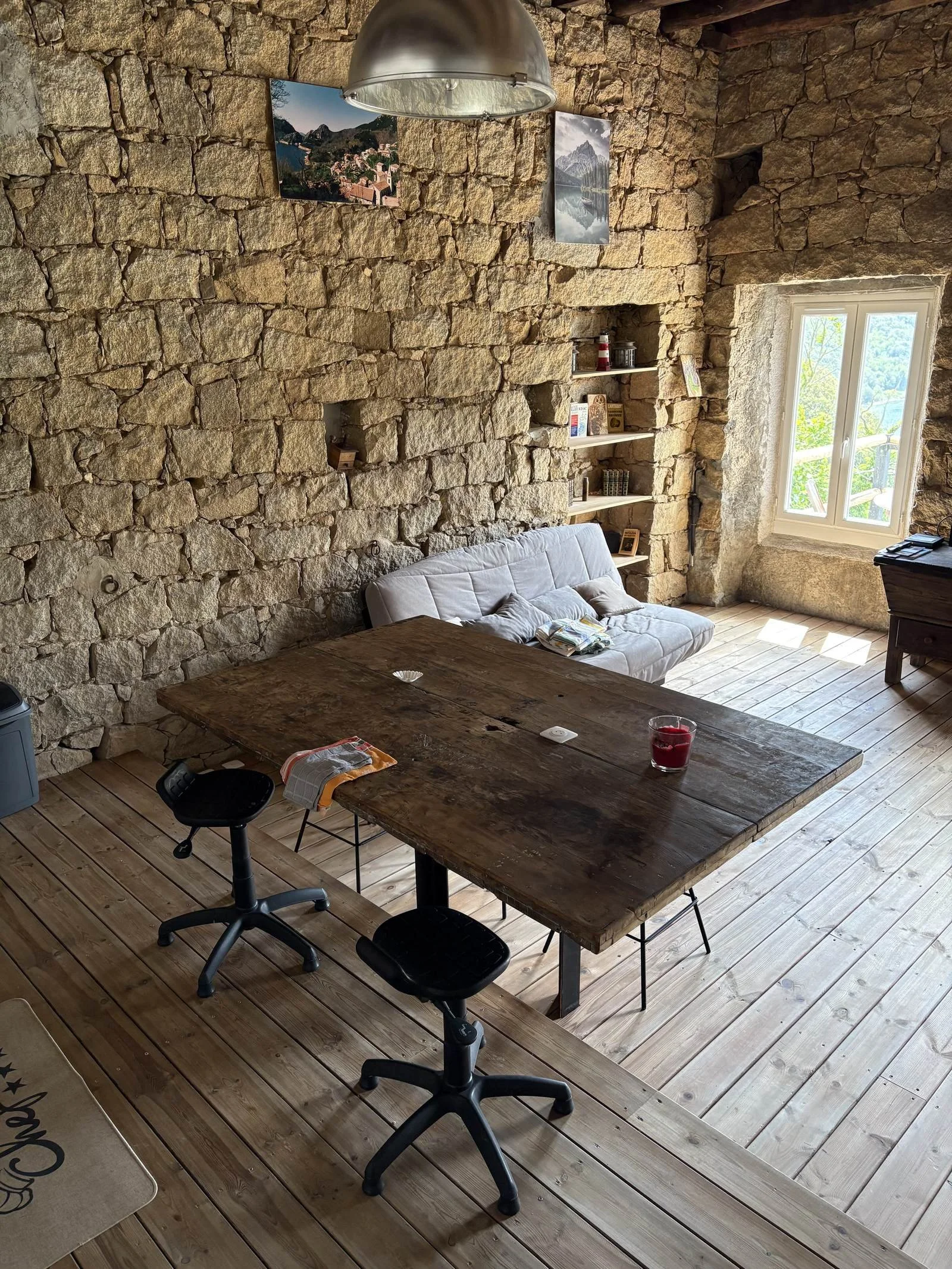 Un intérieur avec un mur en pierre, un canapé blanc, une table en bois massif, des étagères avec des livres, une fenêtre avec vue sur la nature, et une lampe suspendue au plafond.