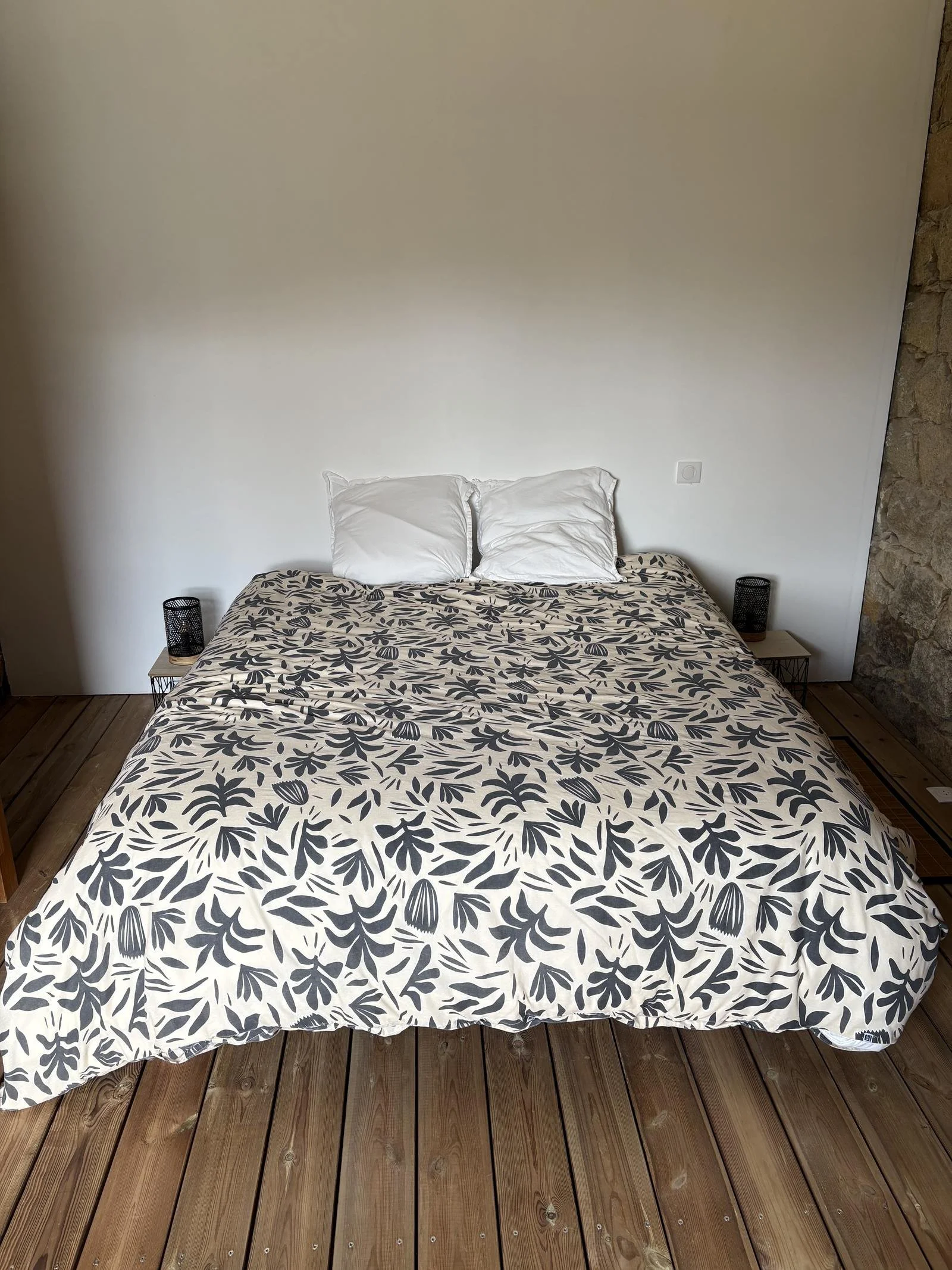 Lit avec une housse à motif floral noir et blanc, deux oreillers blancs, lampes de chevet en métal noir de chaque côté, mur blanc avec prise électrique à droite, sol en bois, mur de pierres sur le côté droit.