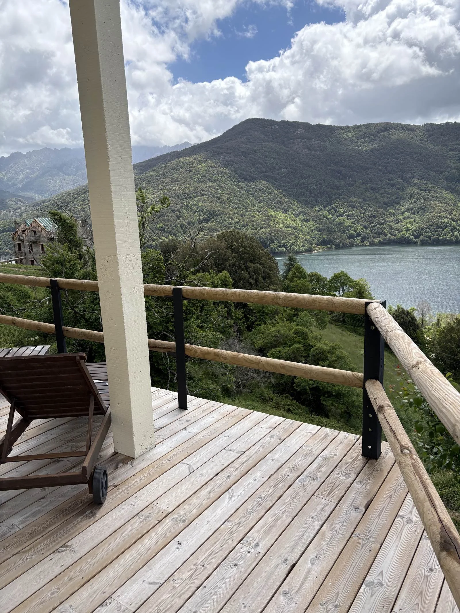 Vue d'une terrasse en bois avec une rambarde en bois et en métal, offrant une vue sur un lac entouré de montagnes verdoyantes. Un fauteuil roulante en bois est placé près de la rambarde.