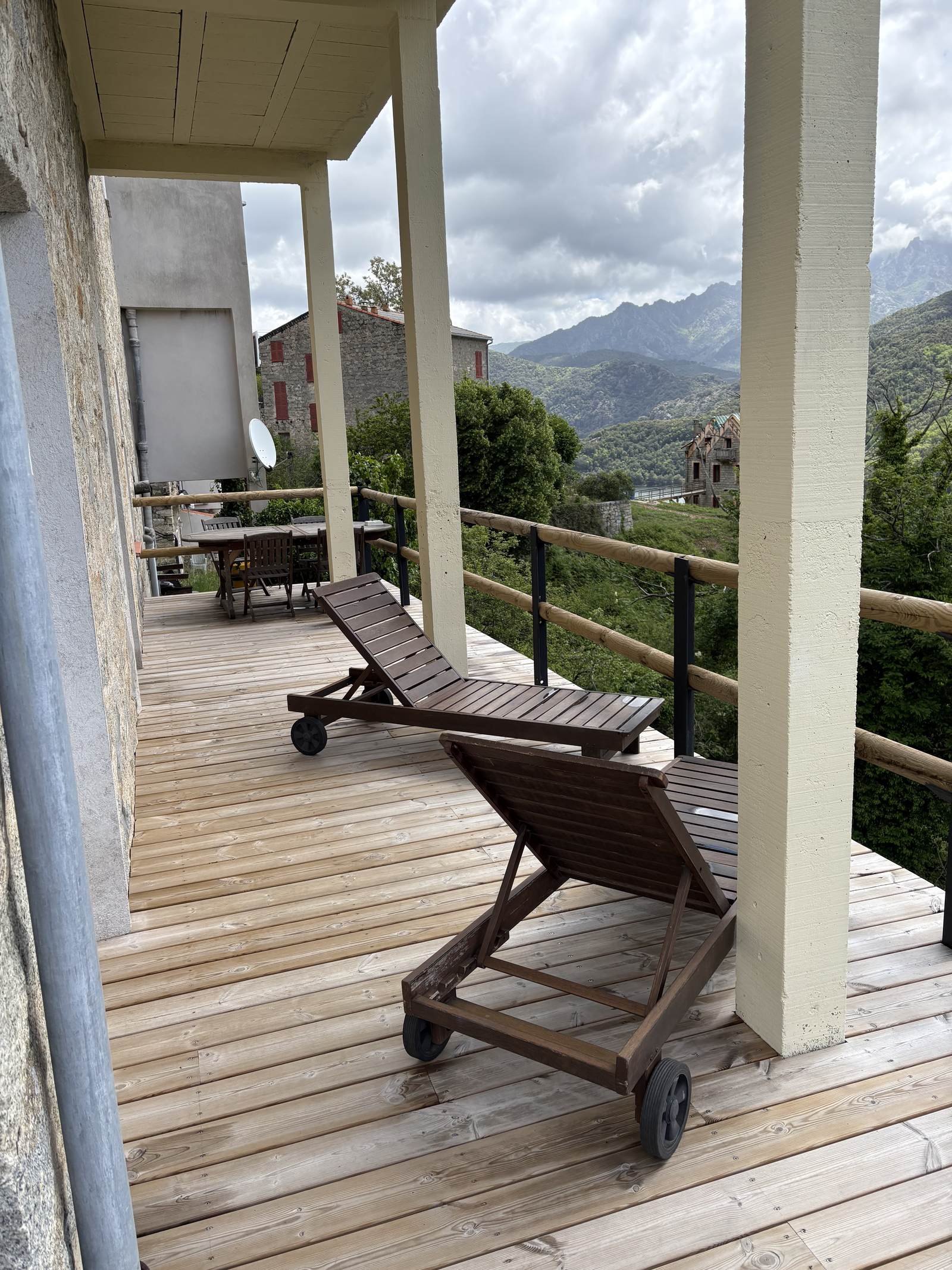 Une terrasse en bois avec des chaises longues en bois et un paysage de montagnes en arrière-plan, sous un ciel nuageux.