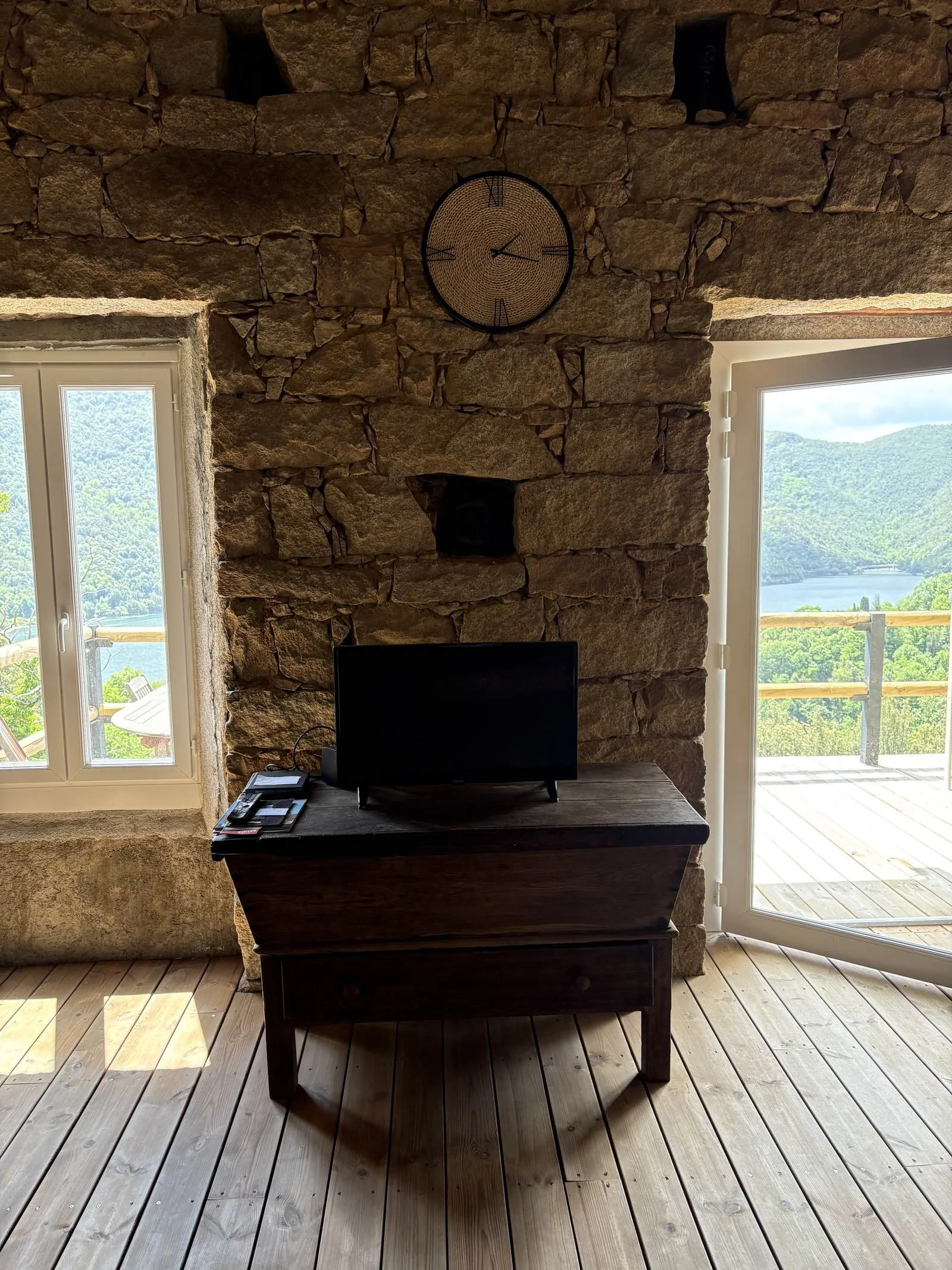 Une pièce avec un mur en pierres, une table en bois sombre, une télévision noir, une horloge ronde, deux fenêtres laissant entrer la lumière naturelle, et un balcon avec une vue sur la nature et un lac au fond.