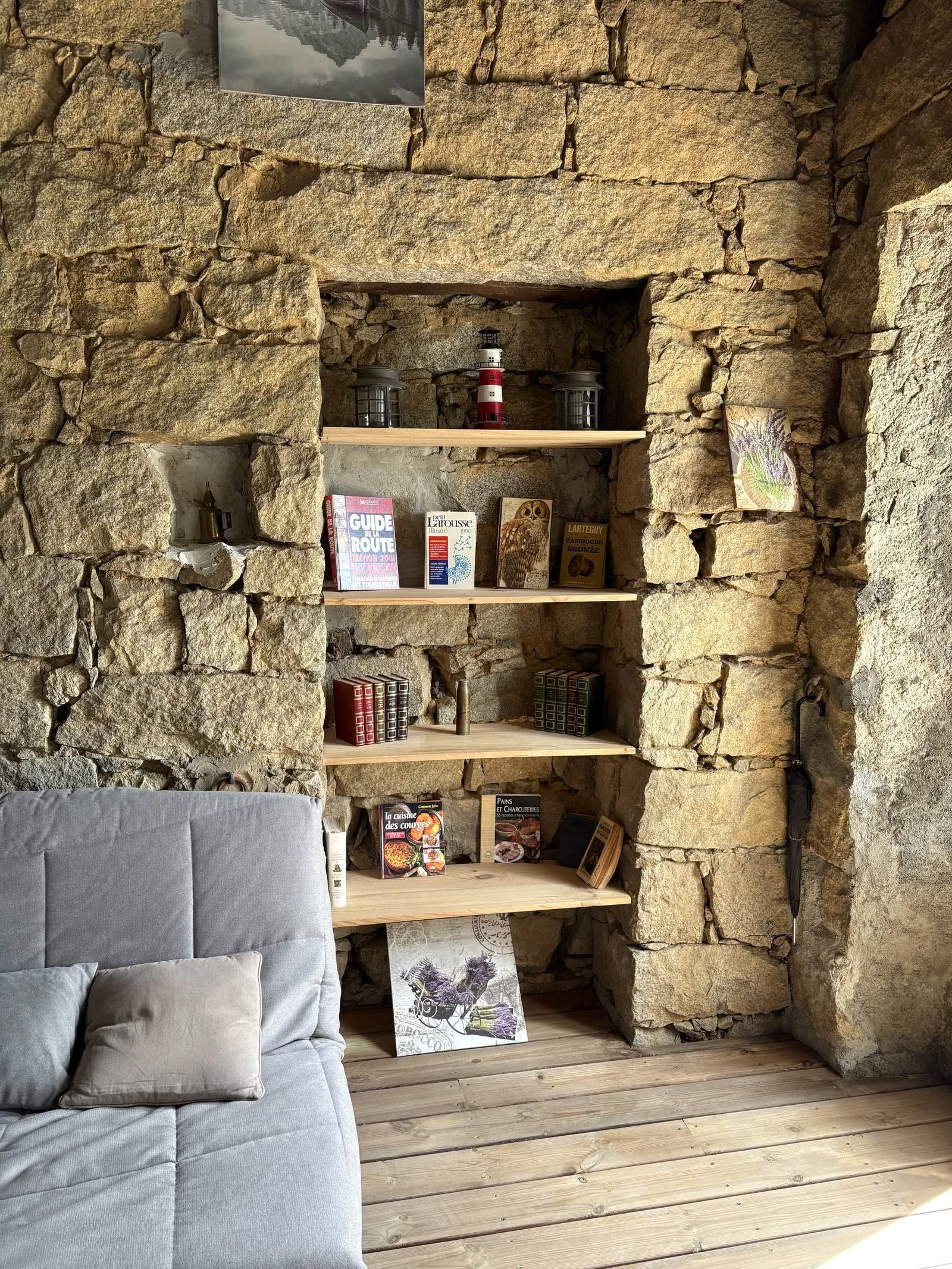 Une étagère en bois intégrée dans un mur de pierres, avec des livres, des illustrations, et des objets décoratifs, partie d'une pièce intérieure avec un lit ou un canapé gris en bas à gauche.