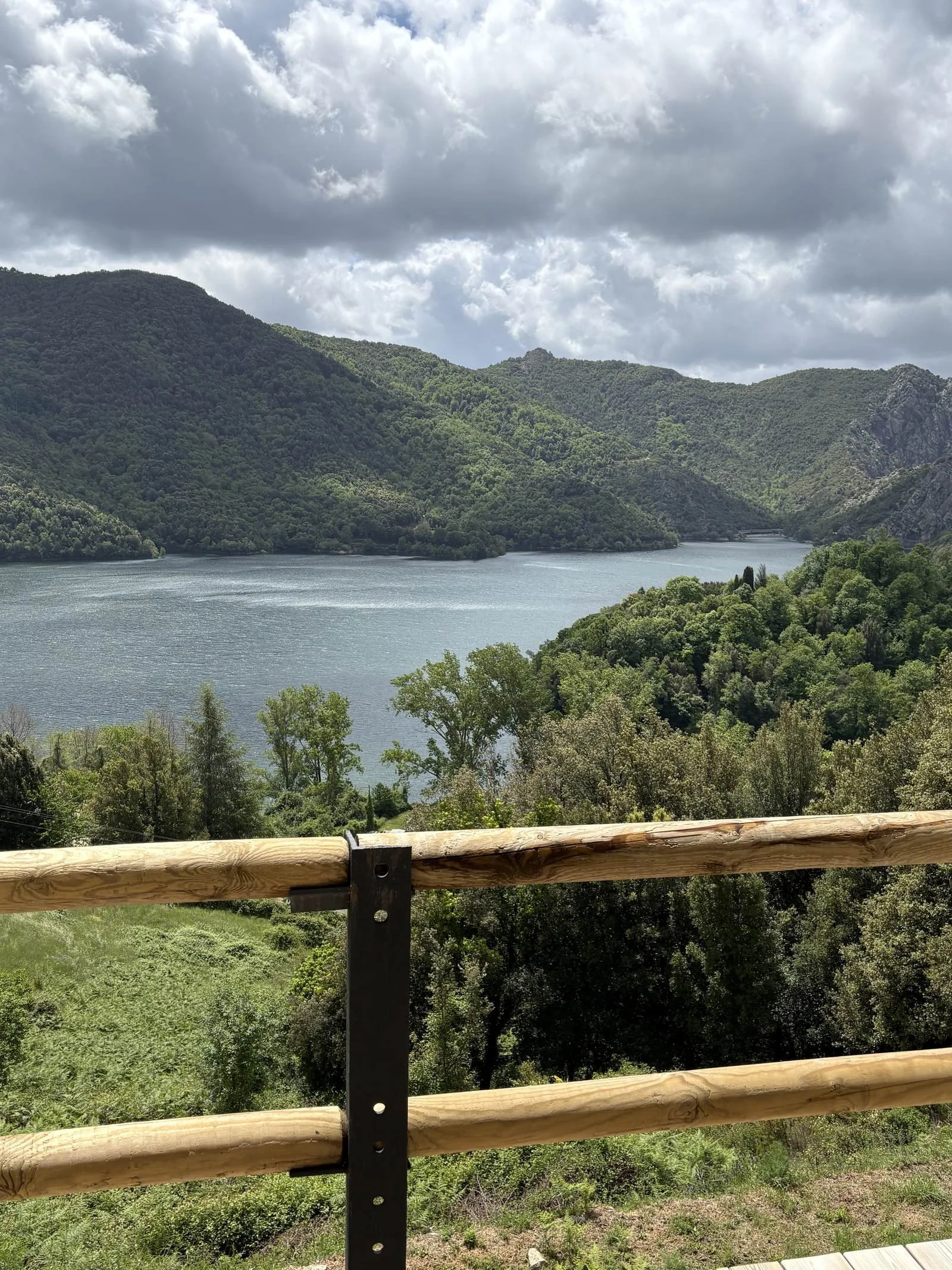 Vue d'une rivière entourée de montagnes verdoyantes sous un ciel nuageux, vue partielle d'une barrière en bois au premier plan.