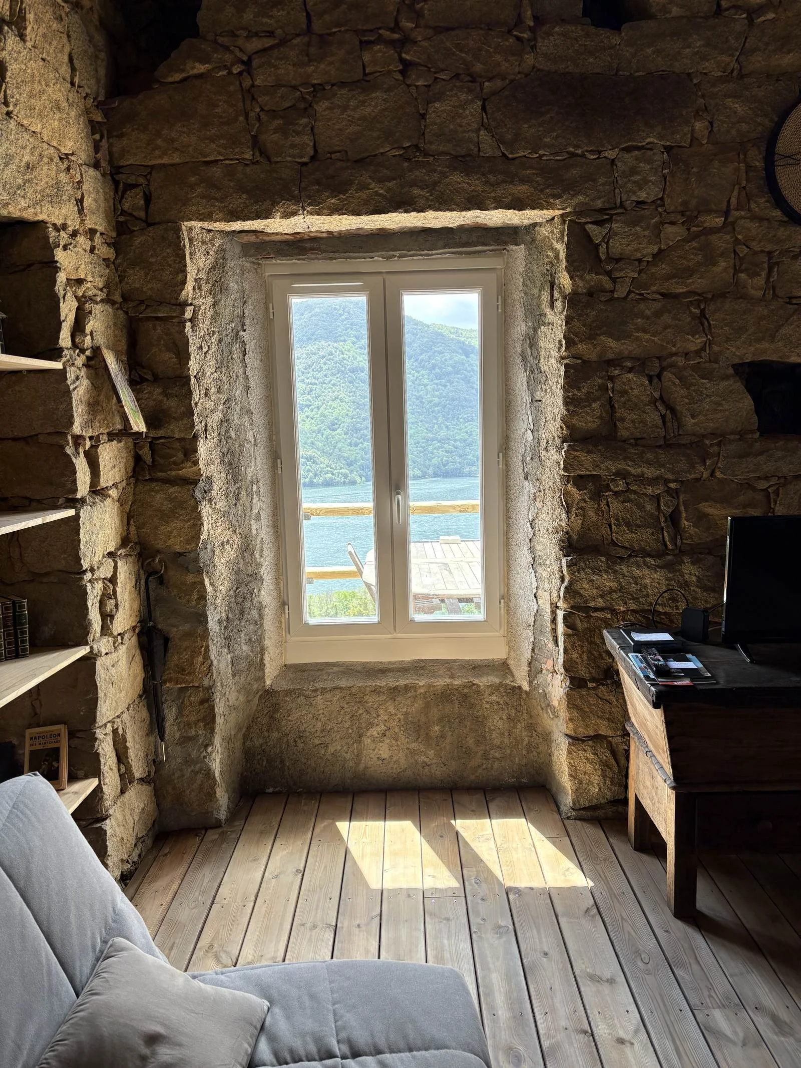 Intérieur d'une pièce en pierre avec une grande fenêtre donnant sur un paysage naturel avec des montagnes et un lac.