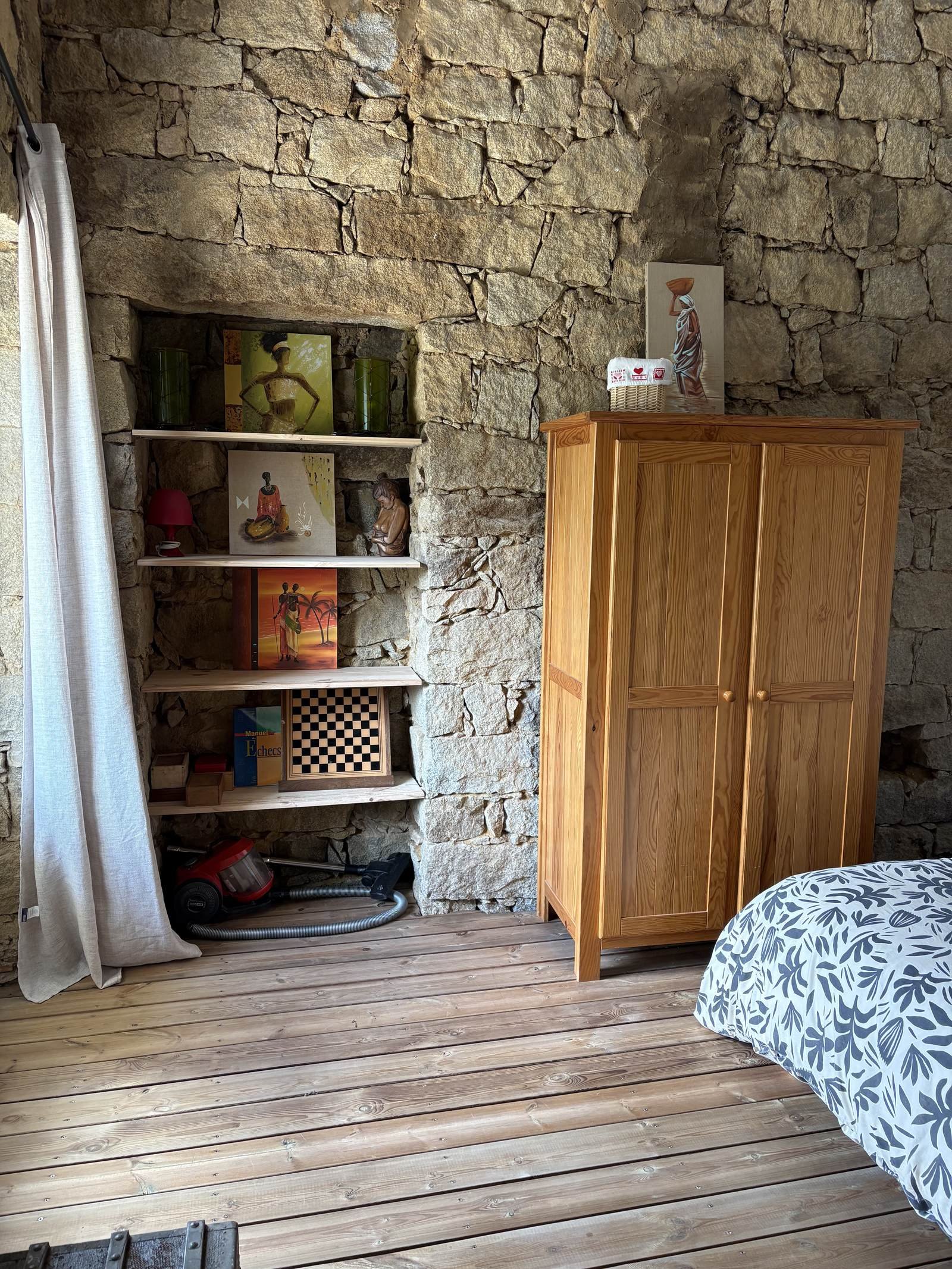 Une pièce avec un mur en pierre, une armoire en bois naturelle, une étagère avec des peintures décoratives, un aspirateur, un rideau blanc et un lit avec une housse à motifs végétaux.