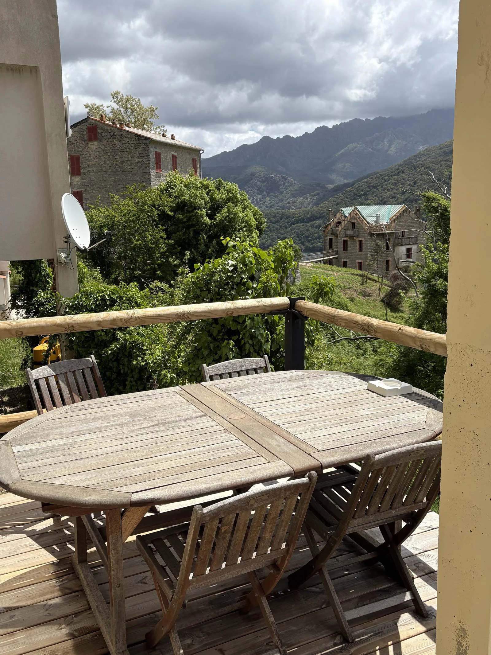 Vue d'une terrasse en bois avec une table en bois entourée de chaises en bois, surplombant un paysage de montagnes et maisons en pierre avec des toits en métal, sous un ciel nuageux.