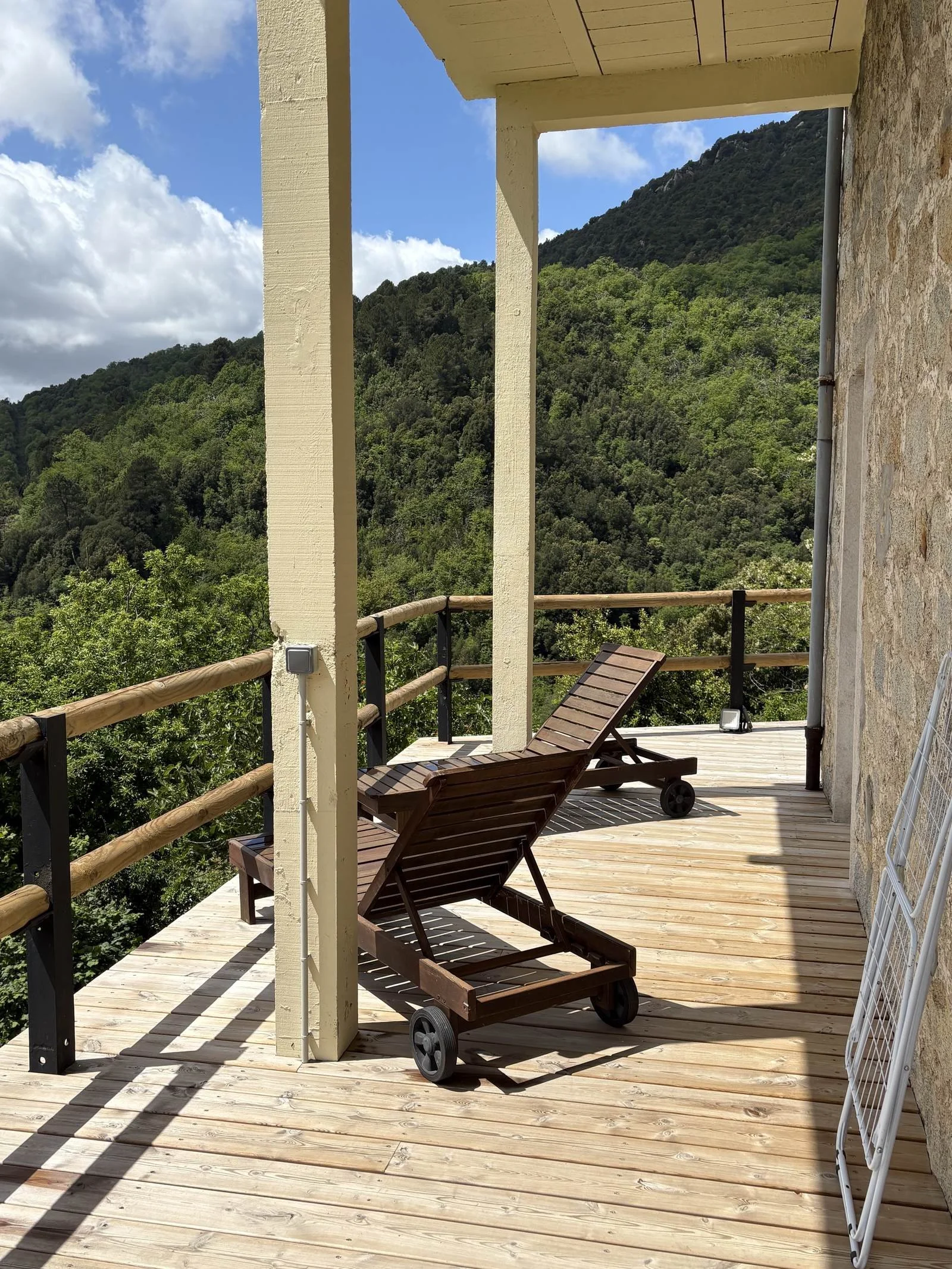 Balcon en bois avec deux fauteuils en bois, entouré de barrières en bois, offrant une vue sur une forêt et des montagnes, avec une partie du mur en pierre, une partie en béton, un éclairage et un panneau en métal