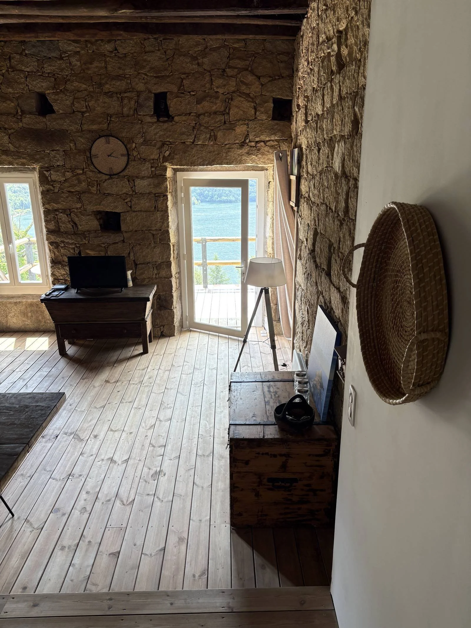 Intérieur d'une maison avec un mur en pierre, une porte-fenêtre donnant sur un lac, une table basse en bois, un lampadaire blanc, une télévision sur une console en bois, un panier en osier accroché au mur, et des tableaux posés sur un meuble en bois.