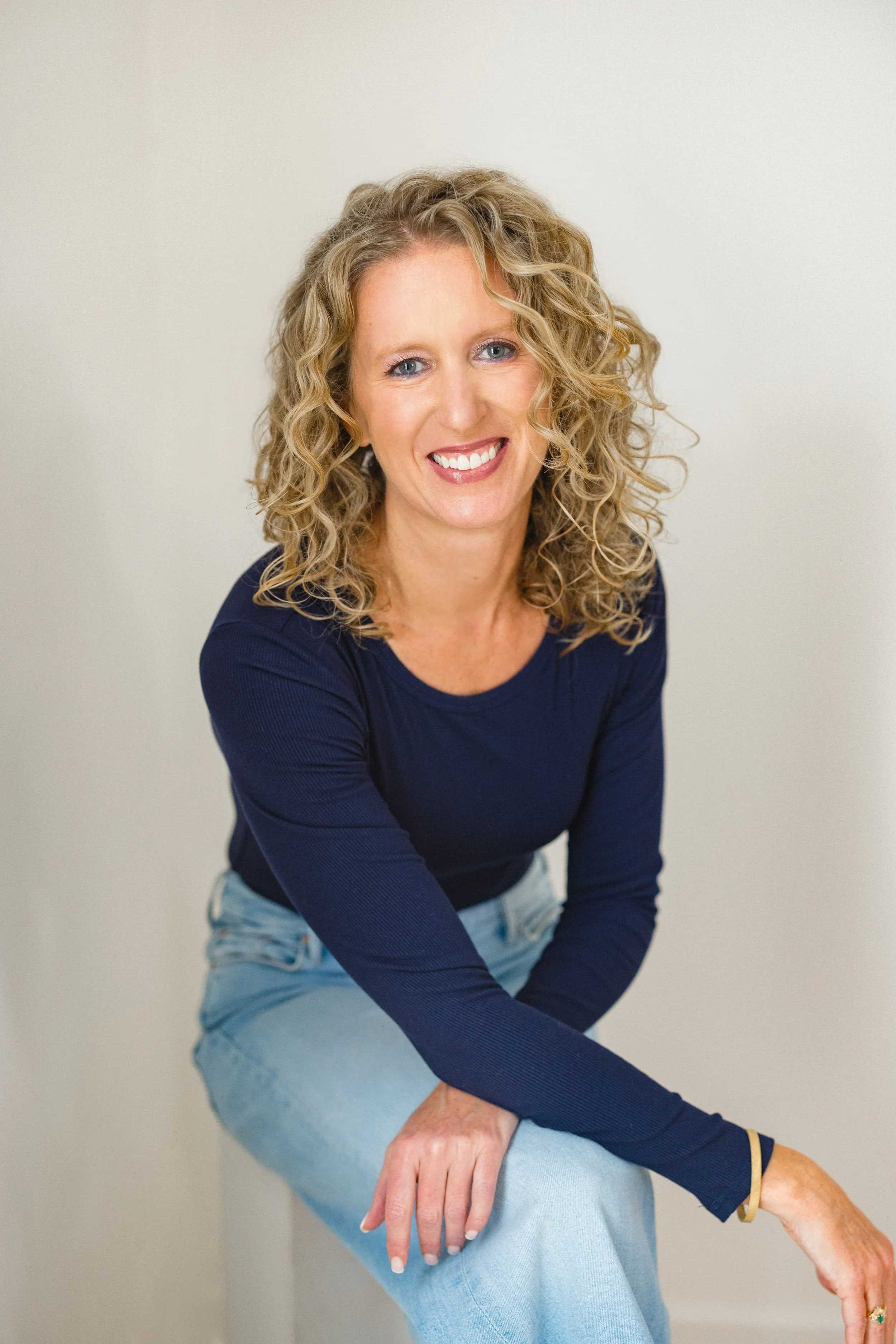 A woman with curly blonde hair wearing a navy blue long-sleeve shirt and light blue jeans, smiling and sitting on a stool against a plain white background.