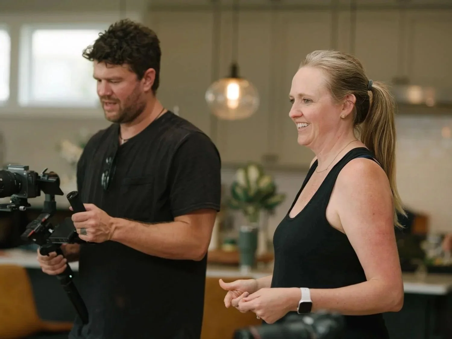 A man with dark, curly hair and a beard is holding a camera stabilizer. A smiling woman with long hair tied back, wearing a black tank top and a smartwatch, is standing next to him in a kitchen setting.