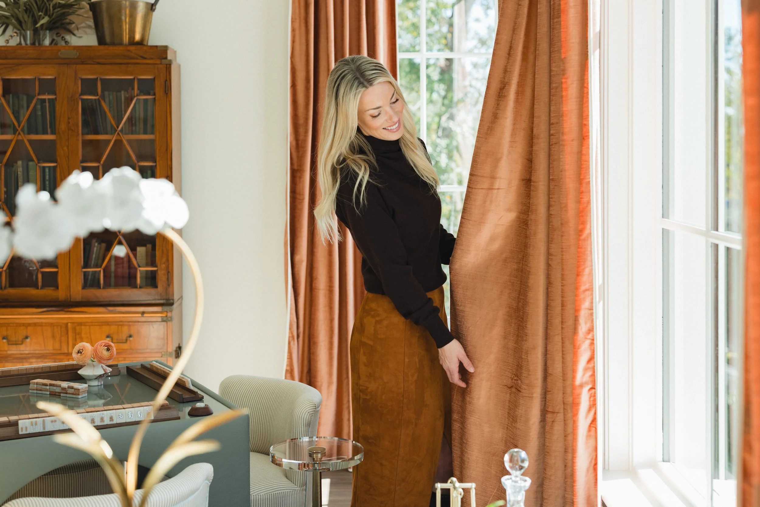 A woman with long blonde hair wearing a black sweater and brown pants smiling while looking outside through orange curtains in a well-lit room.