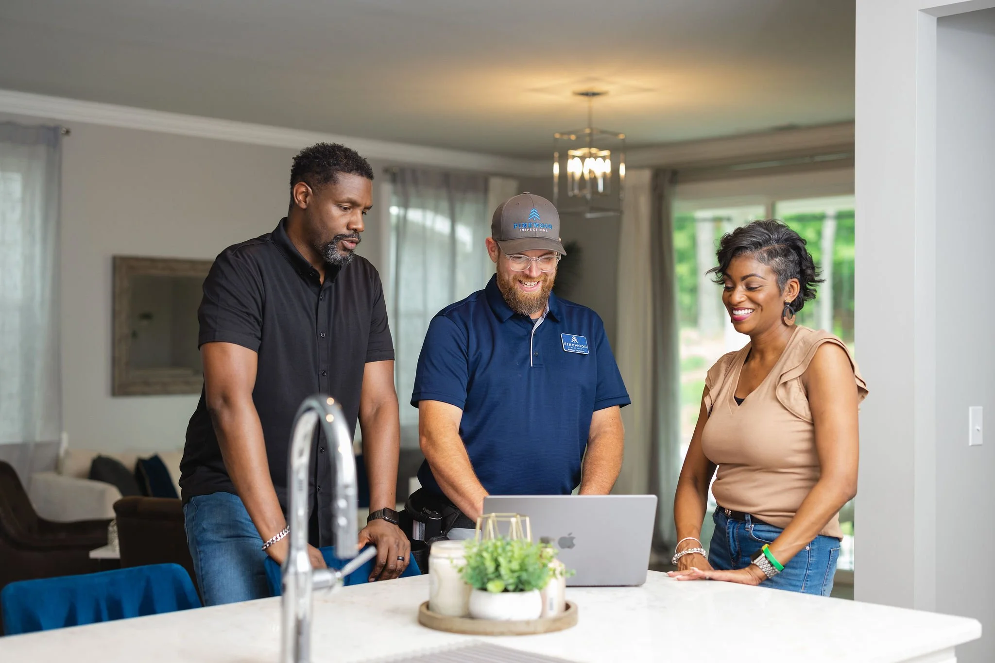 Three people standing around a kitchen island looking at a laptop, smiling, with one person in a service uniform and two in casual clothing.