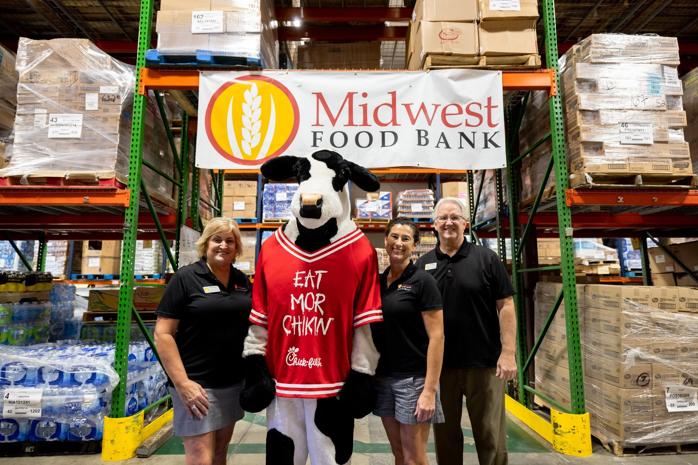 Three people standing with a person in a cow mascot costume inside a warehouse. The mascot is wearing a red jersey with the words "Eat Mor Chikin". Behind them is a sign that reads "Midwest Food Bank" and shelves stocked with boxes and bottled water.