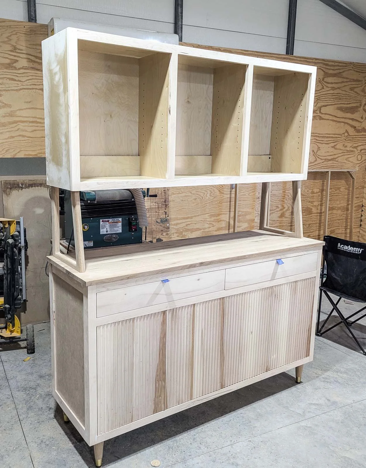 A wooden kitchen cabinet with a countertop and a four-compartment open upper storage unit, set up in a workshop.