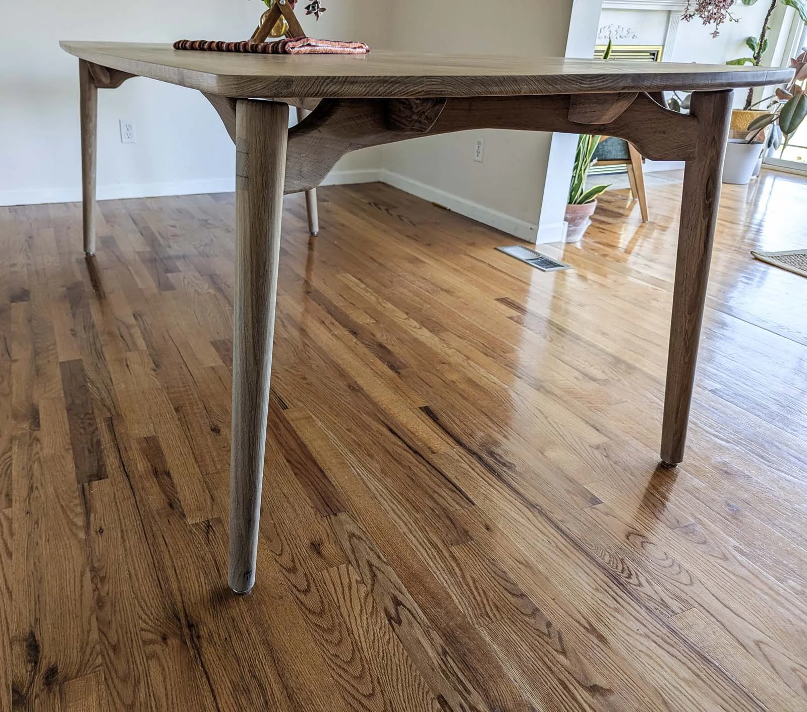 A wooden dining table with a light-colored finish in a room with hardwood flooring, with potted plants and furniture visible in the background.