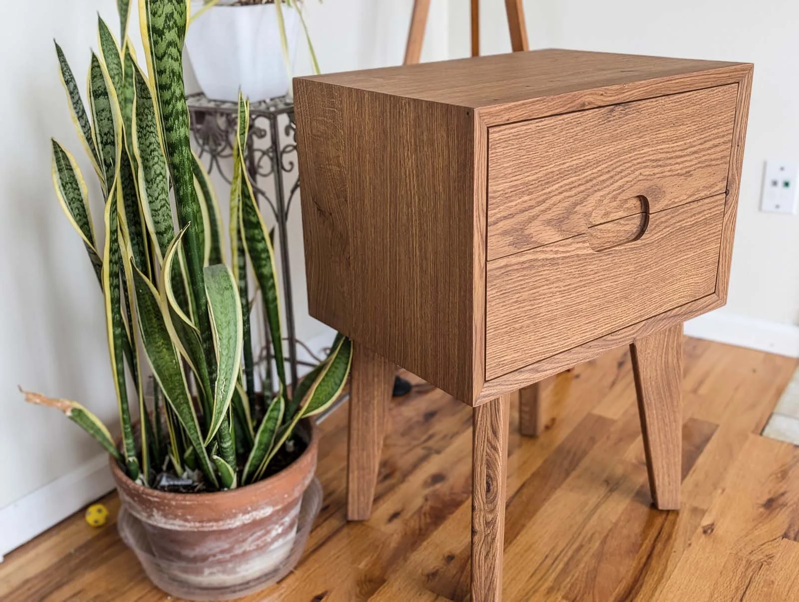 A wooden nightstand or small cabinet with a drawer, standing on four tapered legs, next to a potted snake plant on a hardwood floor.
