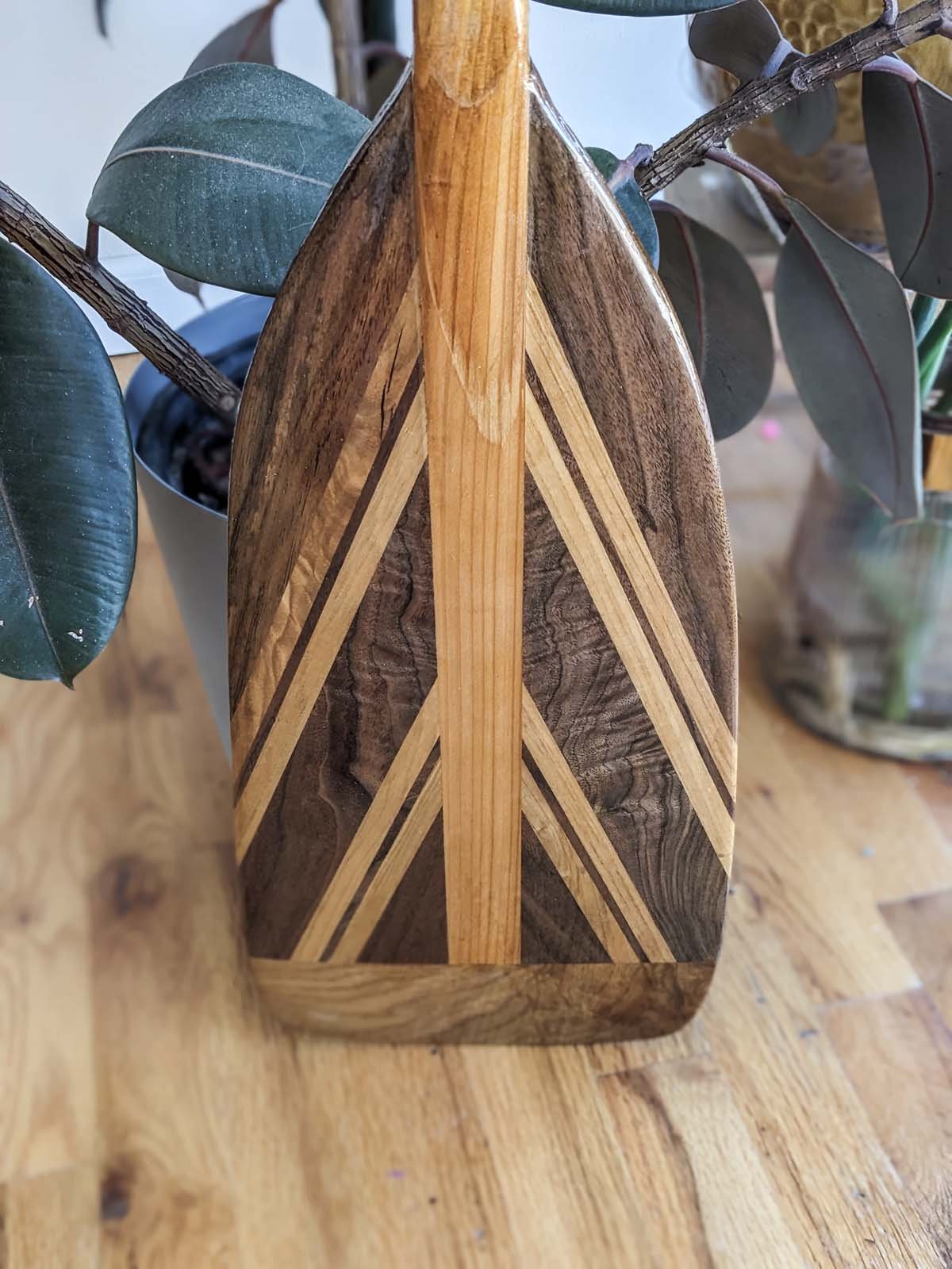 Wooden cutting board with decorative inlay pattern of light and dark wood, placed on a wooden surface with plants nearby.
