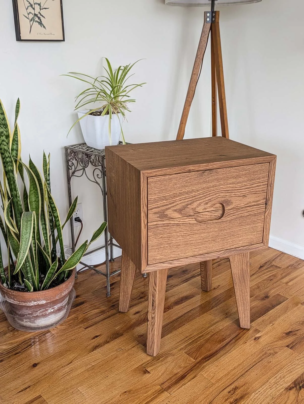 A wooden nightstand with a single drawer in a room with hardwood floors, adjacent to green potted plants and a corner plant stand, with a framed botanical art piece on the wall.