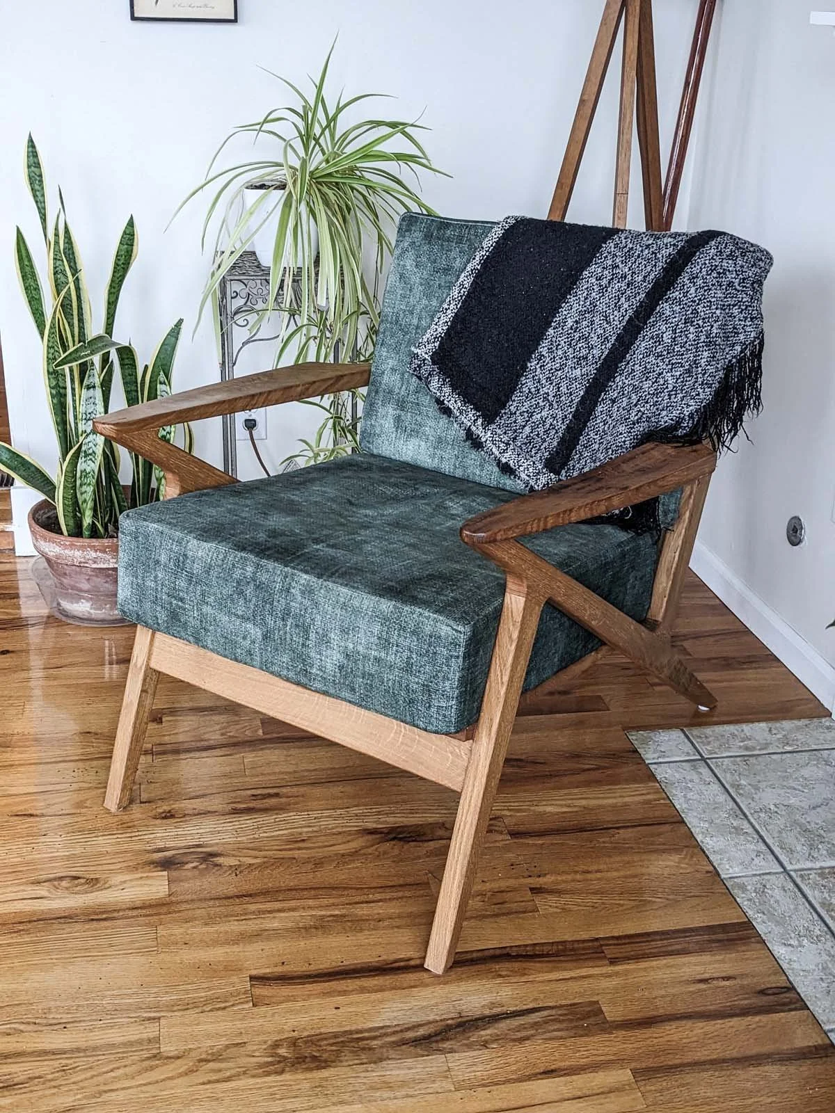 A vintage armchair with teal fabric and wooden arms, with a black and gray throw blanket draped over the back, positioned on a hardwood floor surrounded by houseplants and a small table with a plant.
